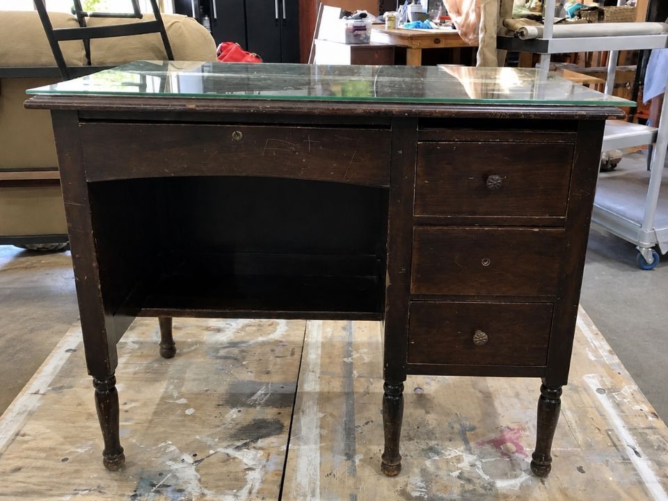 Dark wood antique desk with black top and three drawers, shown in a workshop-like setting.