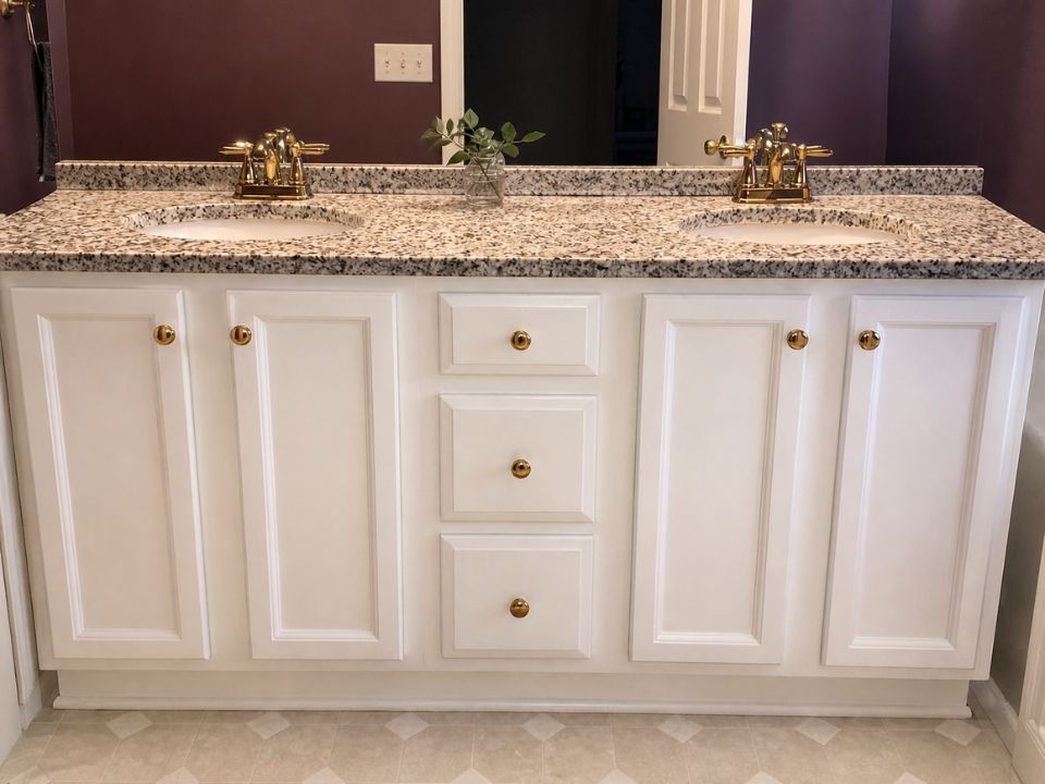 White bathroom vanity with granite countertop, two gold faucets, and central drawers against a purple wall.