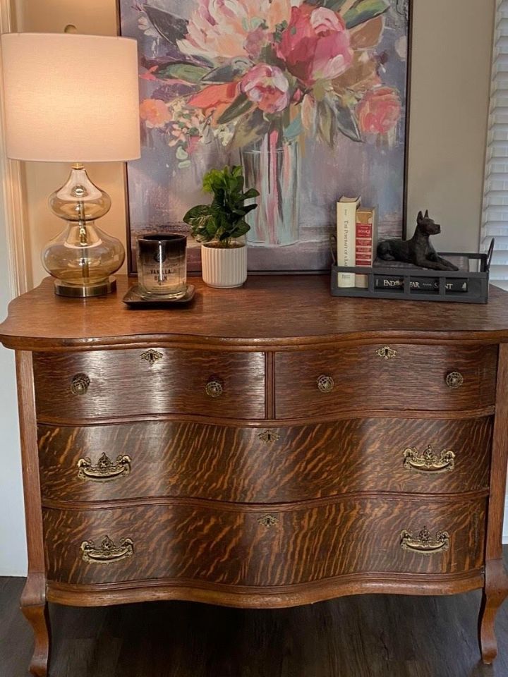 Wooden dresser with lamp, floral painting, small plant, and decor on top in a cozy room.