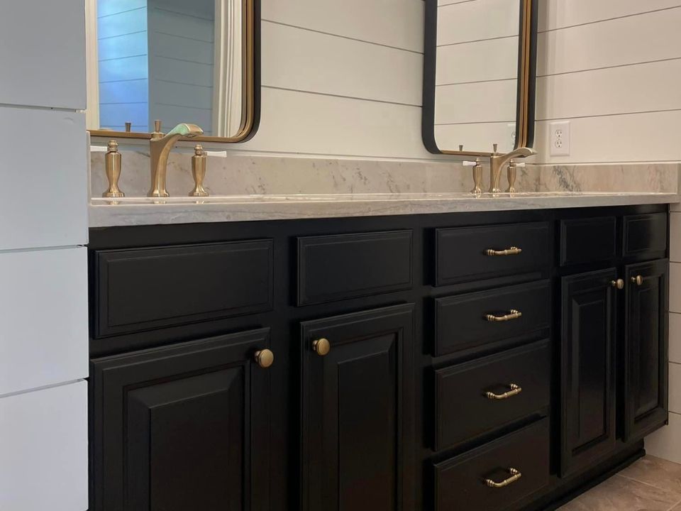 Black bathroom vanity with white countertop and two brass faucets beneath a wall mirror.