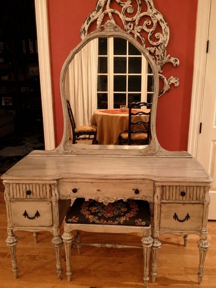 Ornate cream vanity table with mirror against a red wall, with a chair and window reflected in the mirror