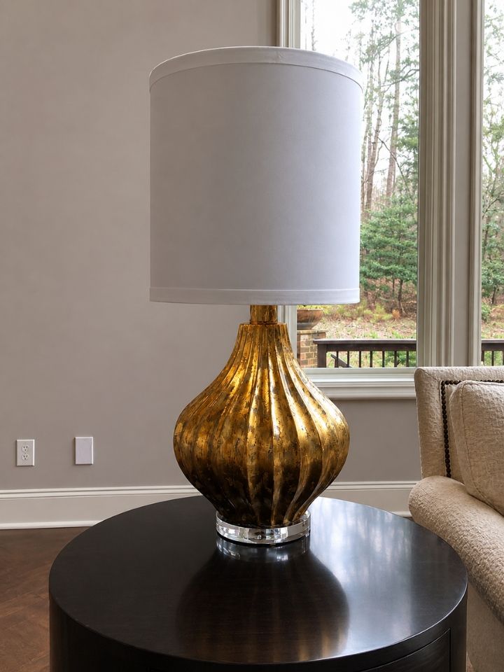 Gold table lamp with white shade on a round black side table near a window