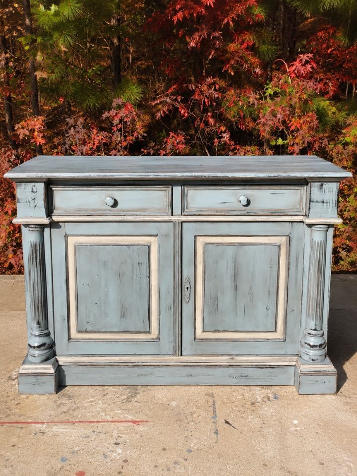 Weathered blue-gray sideboard with drawers and cabinet doors, standing outdoors against autumn foliage.