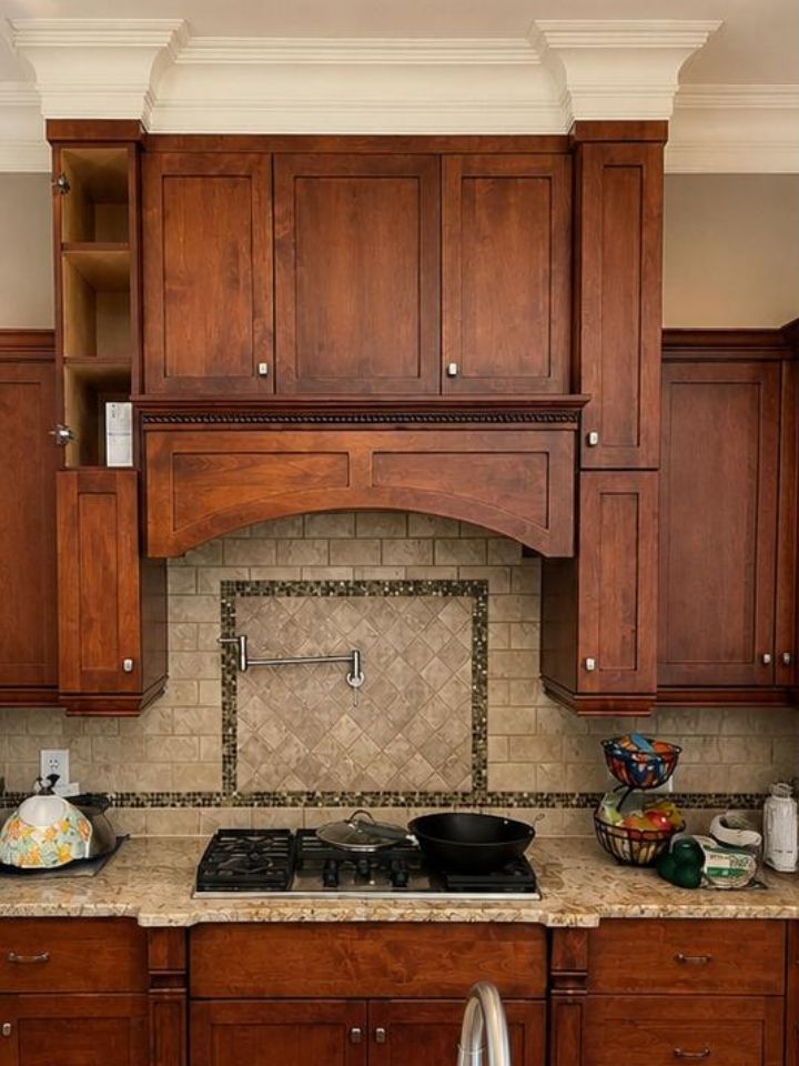 Wooden kitchen cabinets and stove beneath a tiled range hood with granite countertop and decorative items.