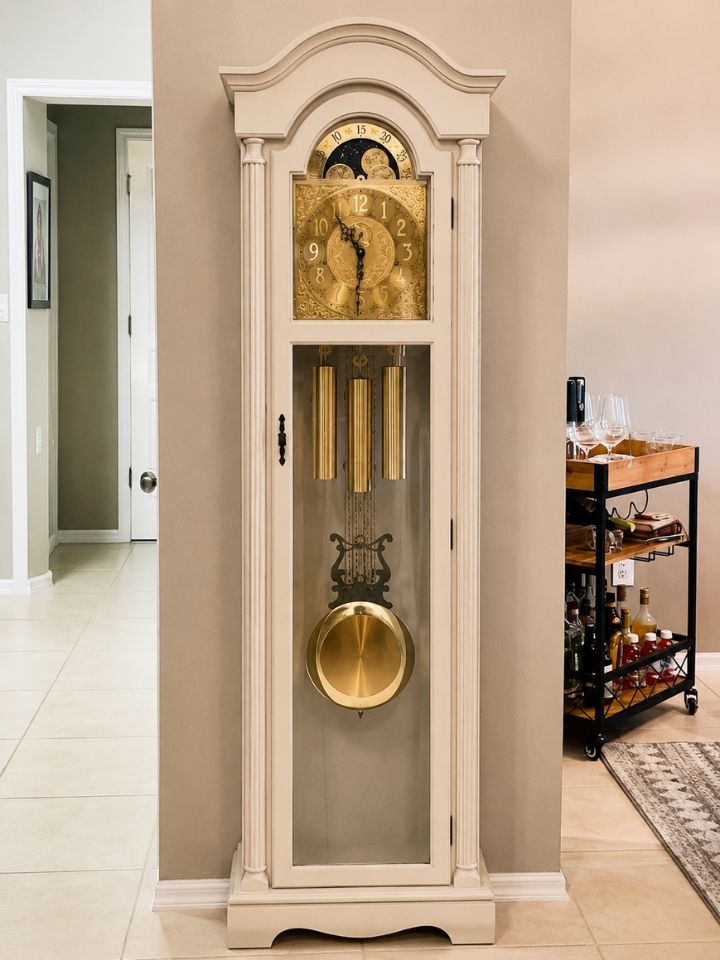 Tall cream grandfather clock with gold face and pendulum in a hallway with white tile floor