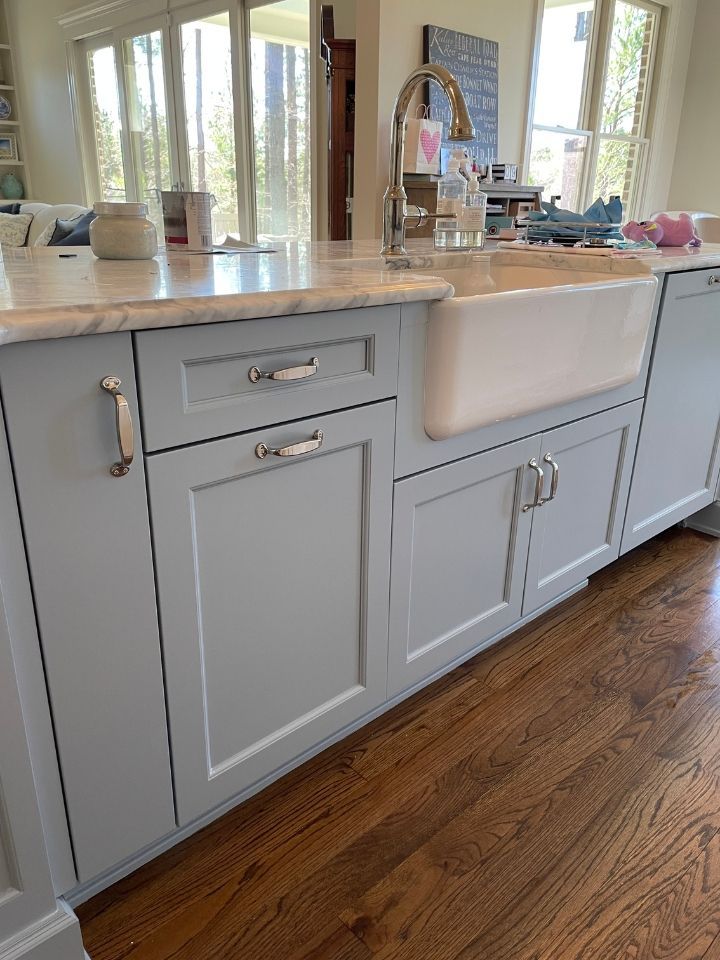 Light gray farmhouse kitchen sink cabinet with white countertop, brass faucet, and hardwood floor