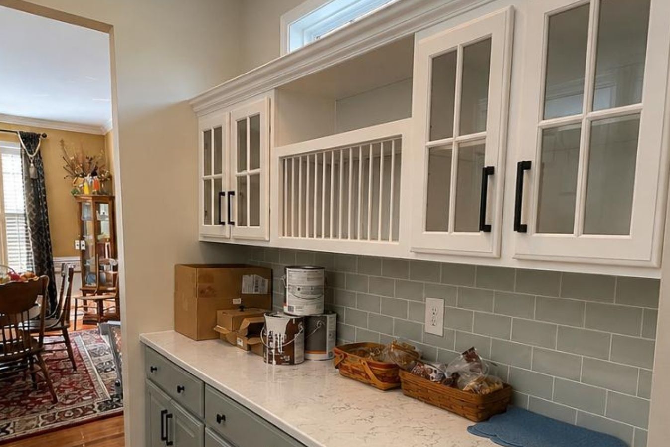 White kitchen cabinets with glass doors and pale green backsplash above a countertop with baskets and jars.
