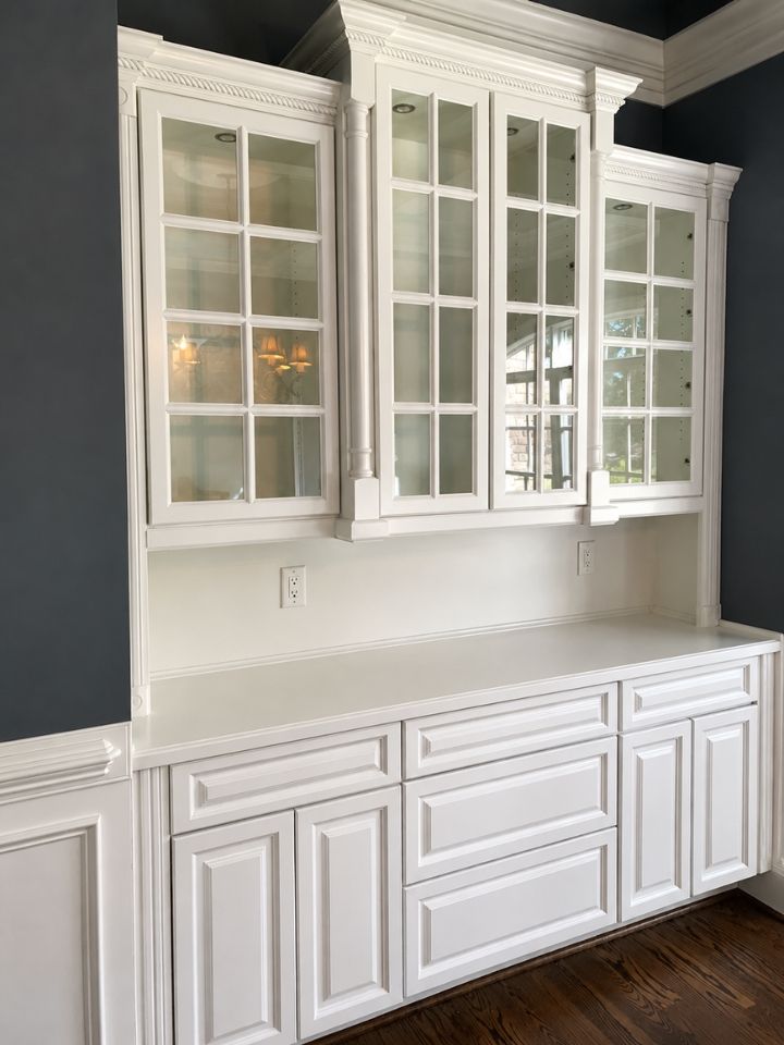White built-in cabinets with glass-front upper doors and drawers against a gray wall, under decorative molding.