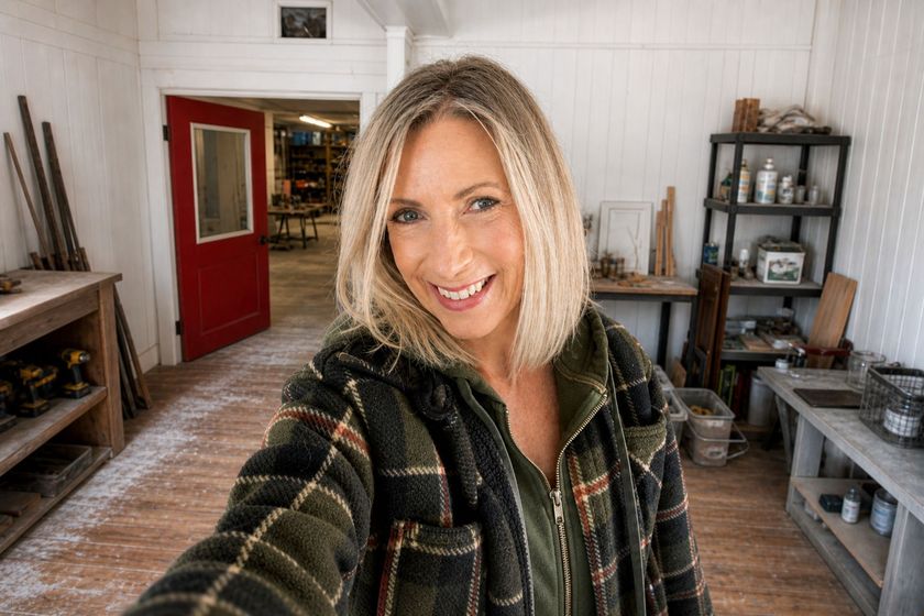 Selfie in a rustic room with wood floors, shelves, and a red door; smiling woman in a plaid jacket.