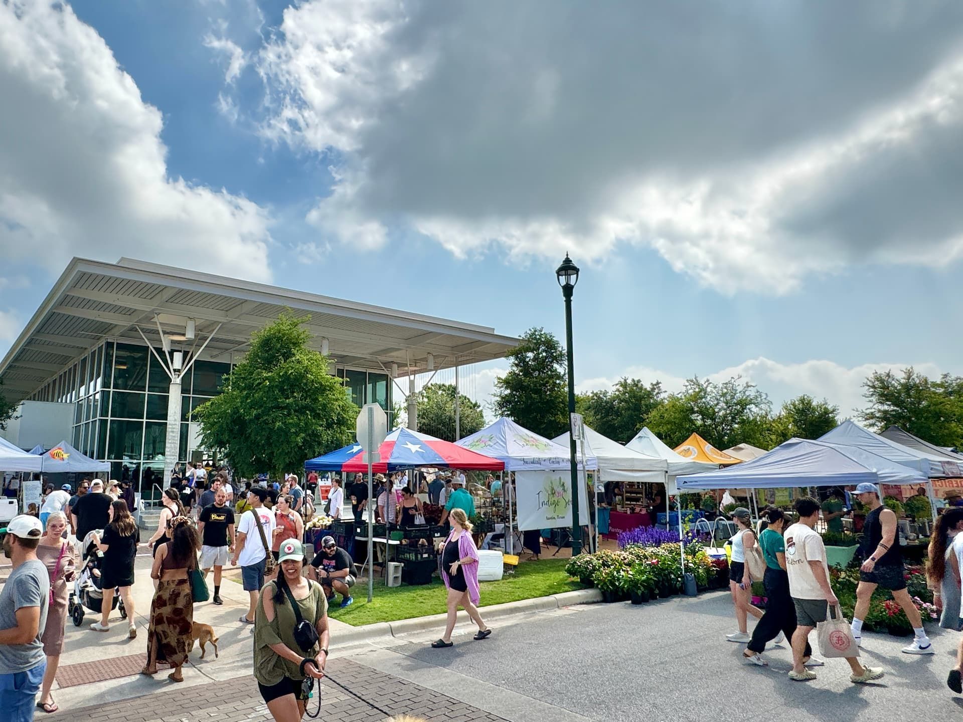 Outdoor farmers market with people, vendors, tents, and a modern building under a partly cloudy sky.