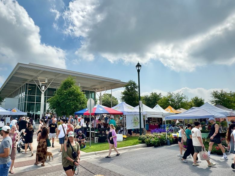Outdoor farmers market with people, vendors, tents, and a modern building under a partly cloudy sky.