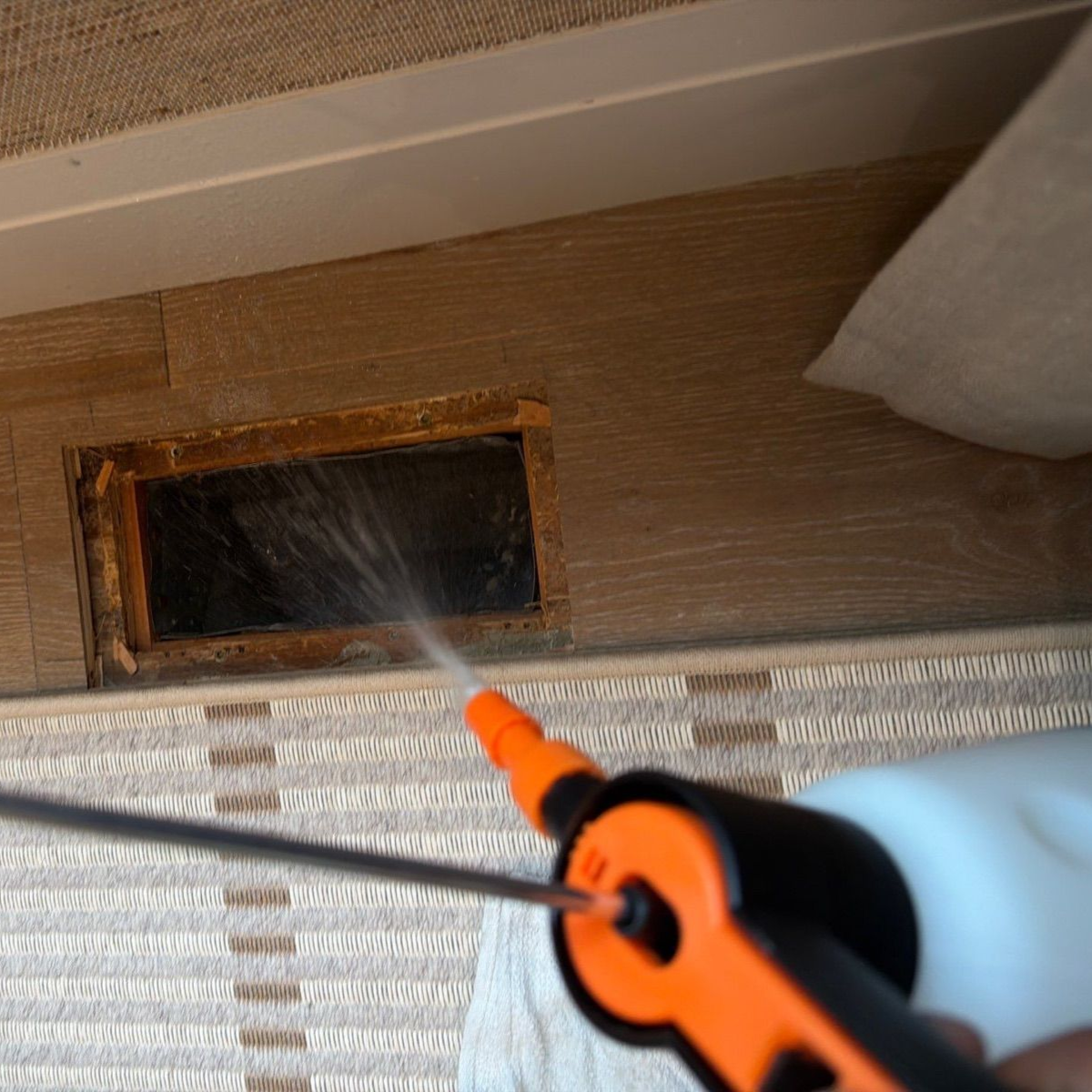 Orange spray nozzle cleaning a dark vent opening under a beige surface