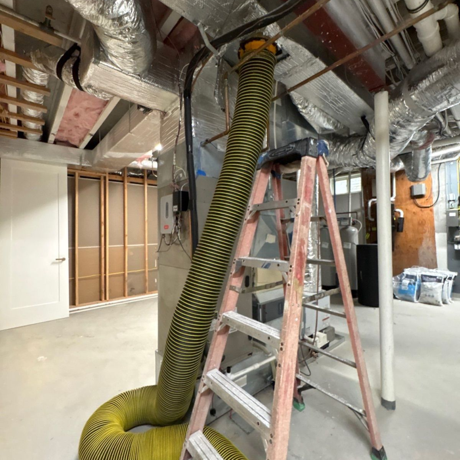 Yellow-and-black striped flexible duct hanging from a ladder in an unfinished room with exposed ceiling insulation.