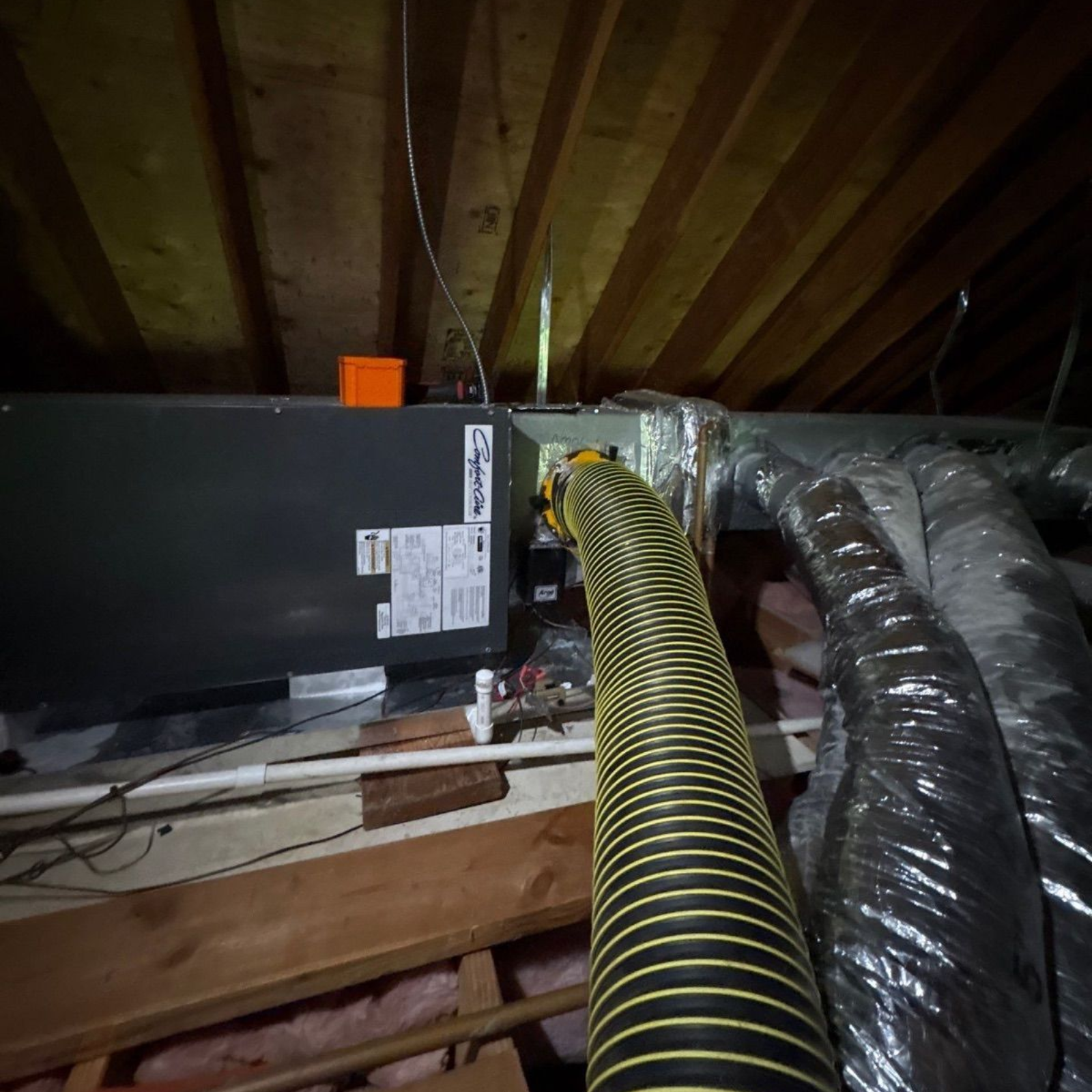Attic crawlspace with black HVAC ducting, exposed wooden rafters, and insulated pipes running overhead
