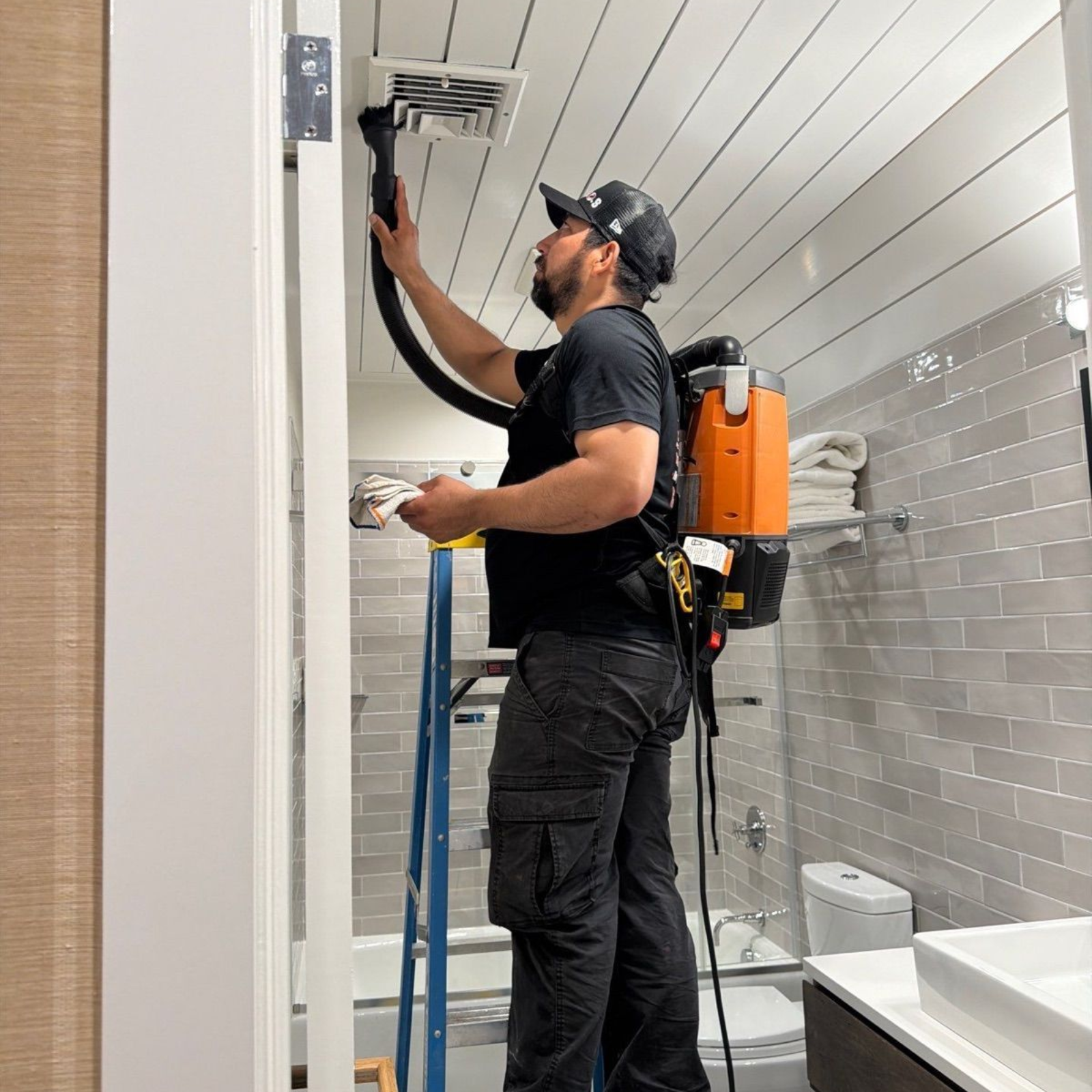 Technician using a spray wand while standing on a ladder in a tiled room, wearing an orange tank sprayer.