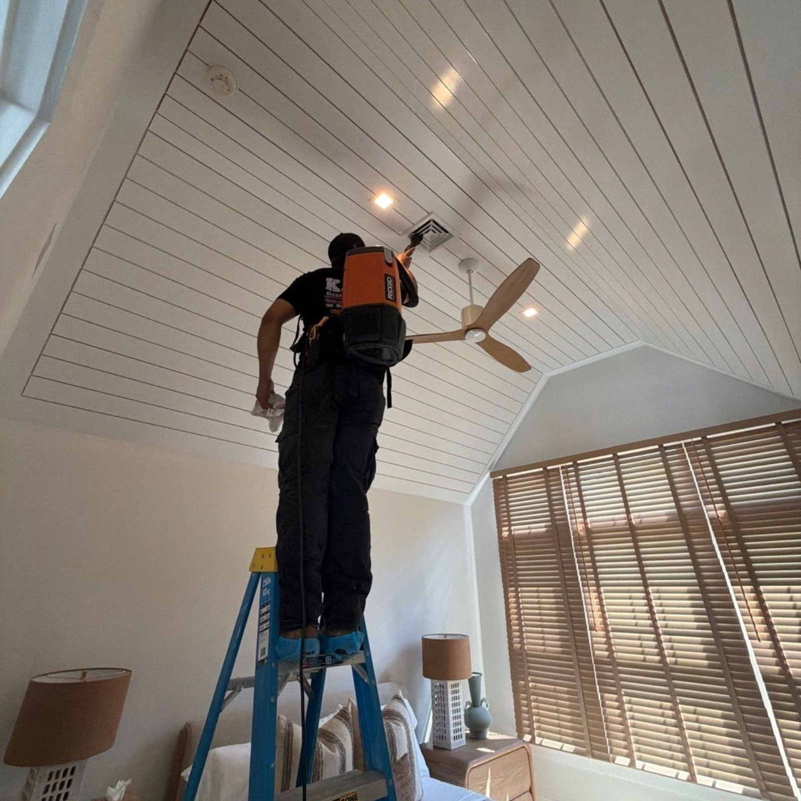 Person on a blue ladder painting a white ceiling in a bright room with blinds.