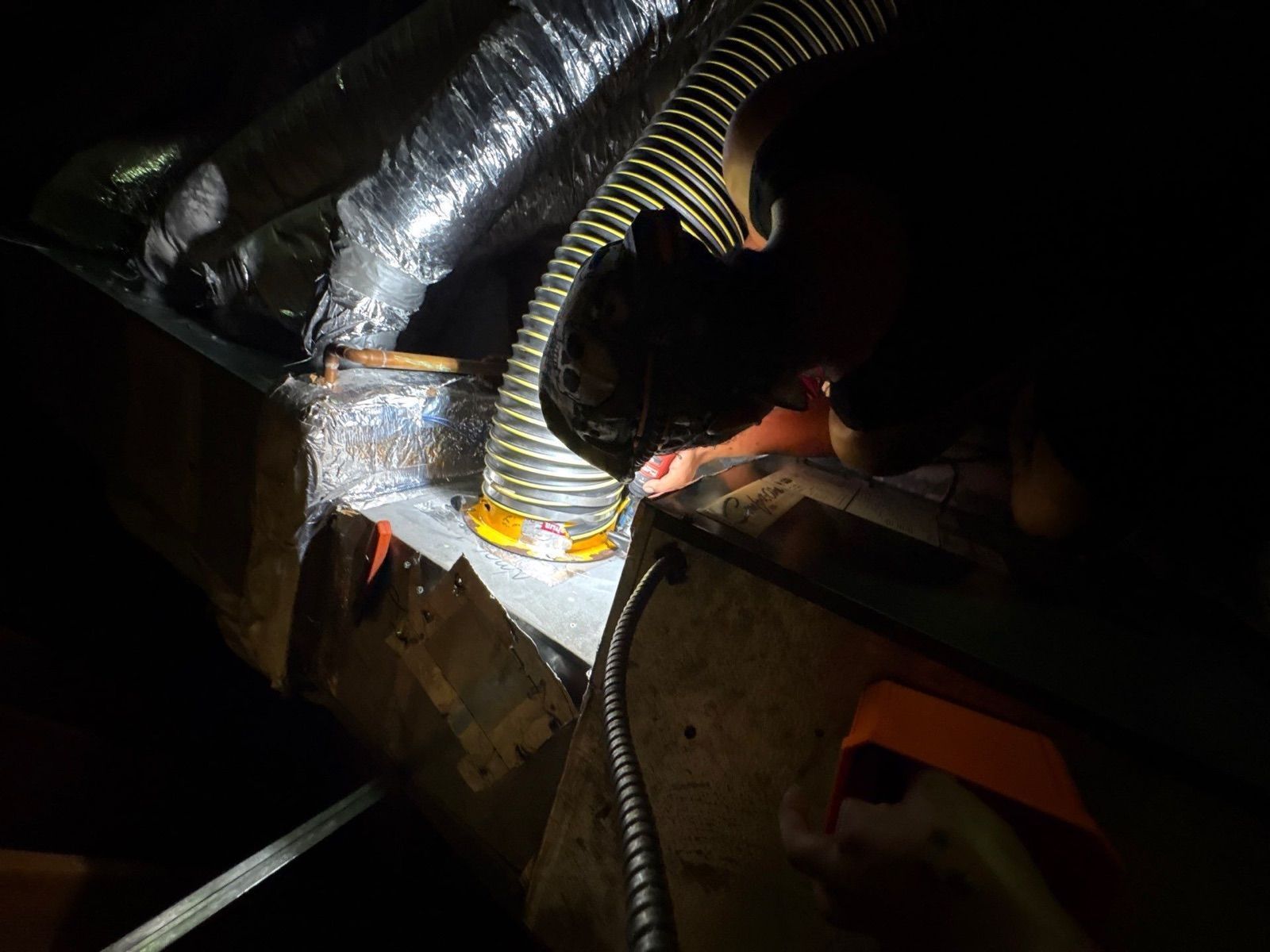 Person working inside a dark crawl space with ductwork, using a flashlight on insulated pipes.