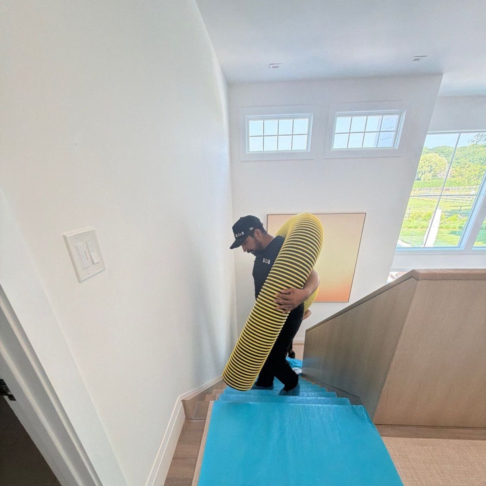 Person carrying a striped mattress up a stairway with a blue protective runner in a hallway.