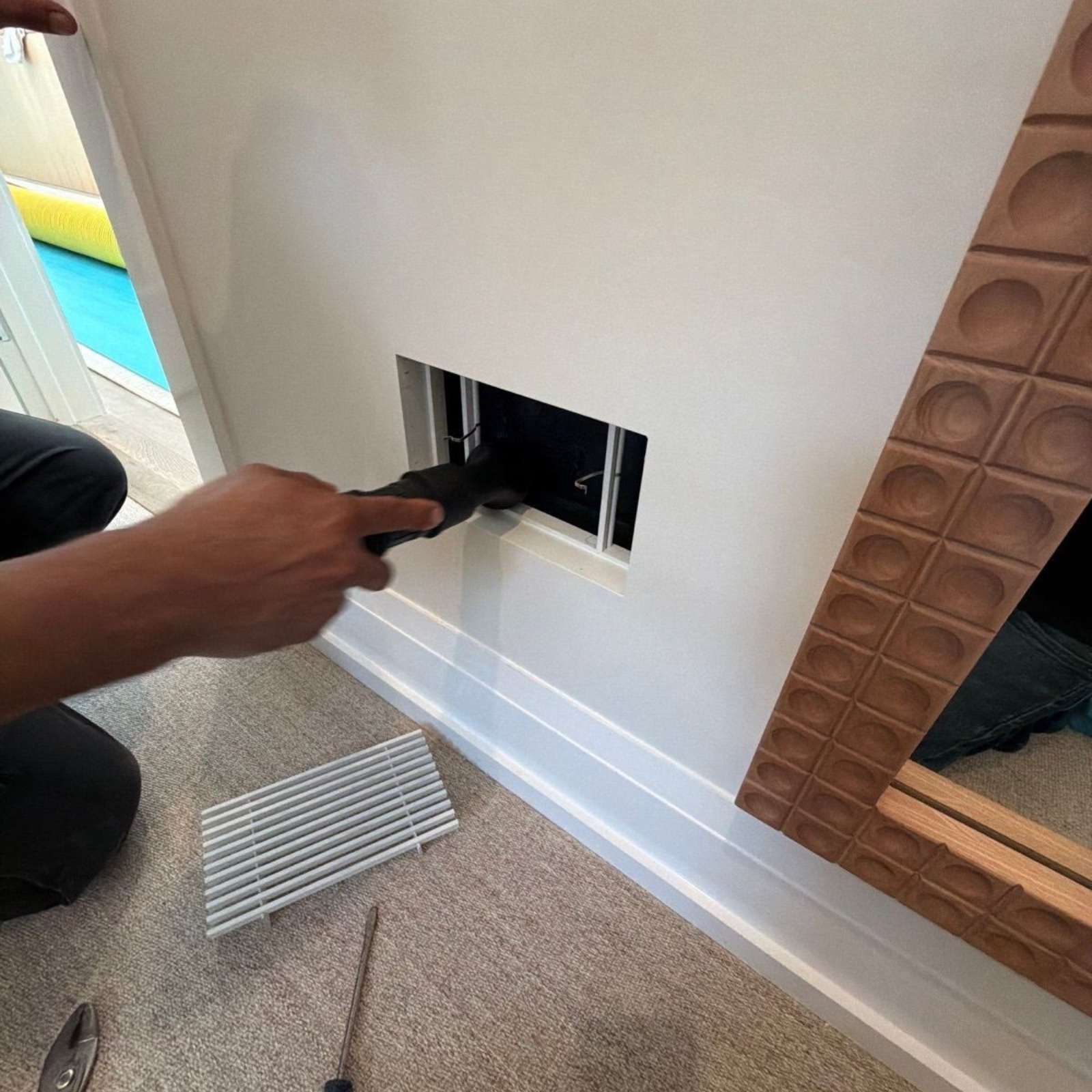 Hand inserting a black cable into a wall outlet opening beside a vent on a carpeted floor
