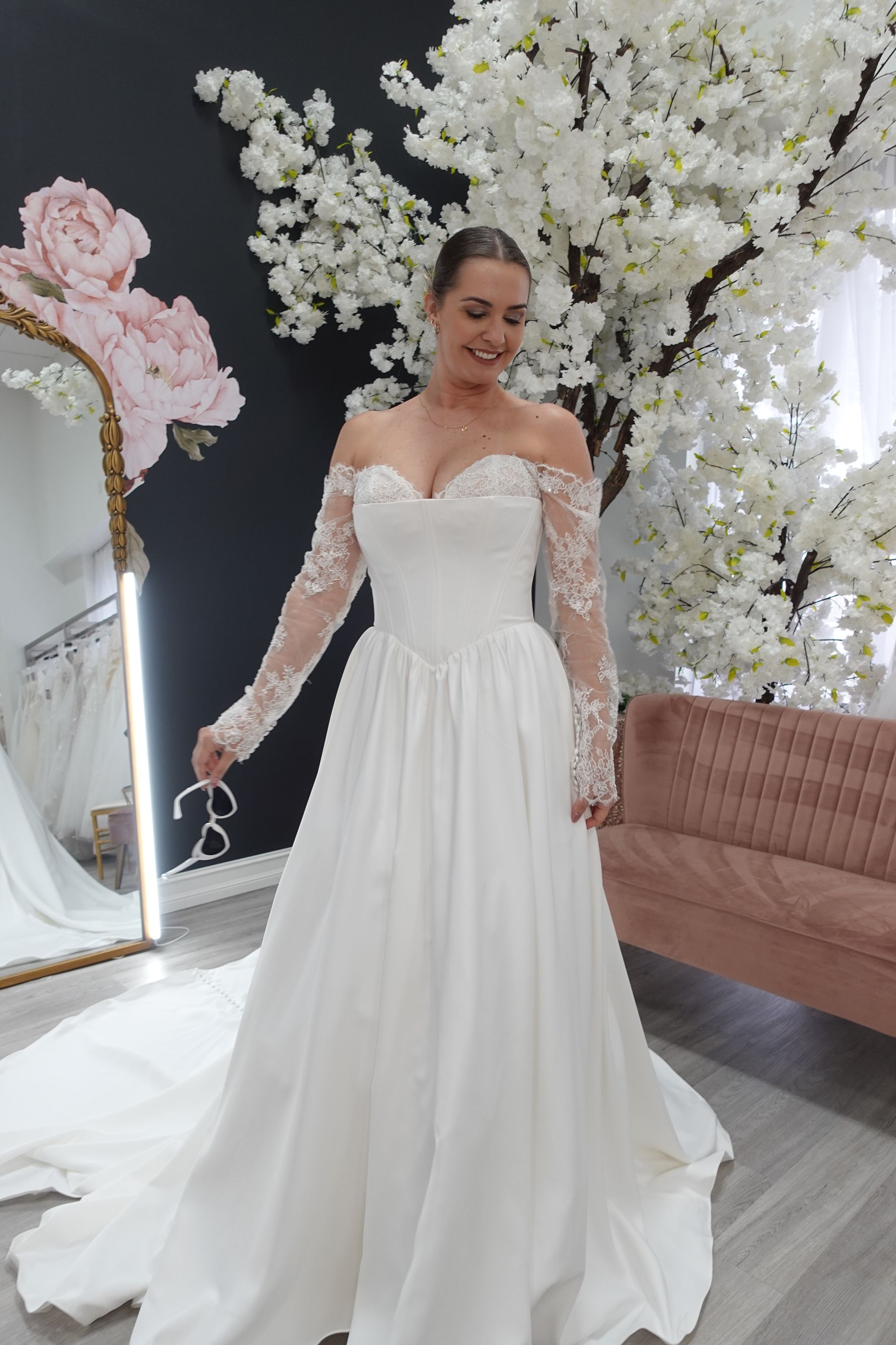 Woman in a white off-the-shoulder wedding dress smiles in a bridal shop; cherry blossom backdrop.