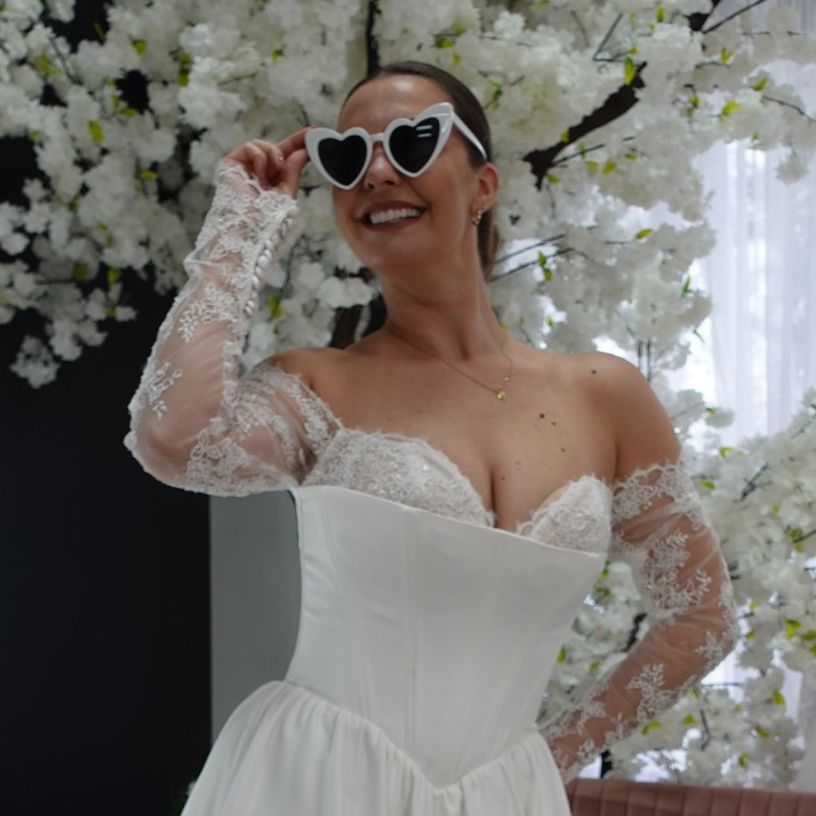 Woman wearing heart-shaped sunglasses, a white dress, and lace gloves smiles in front of a flower backdrop.