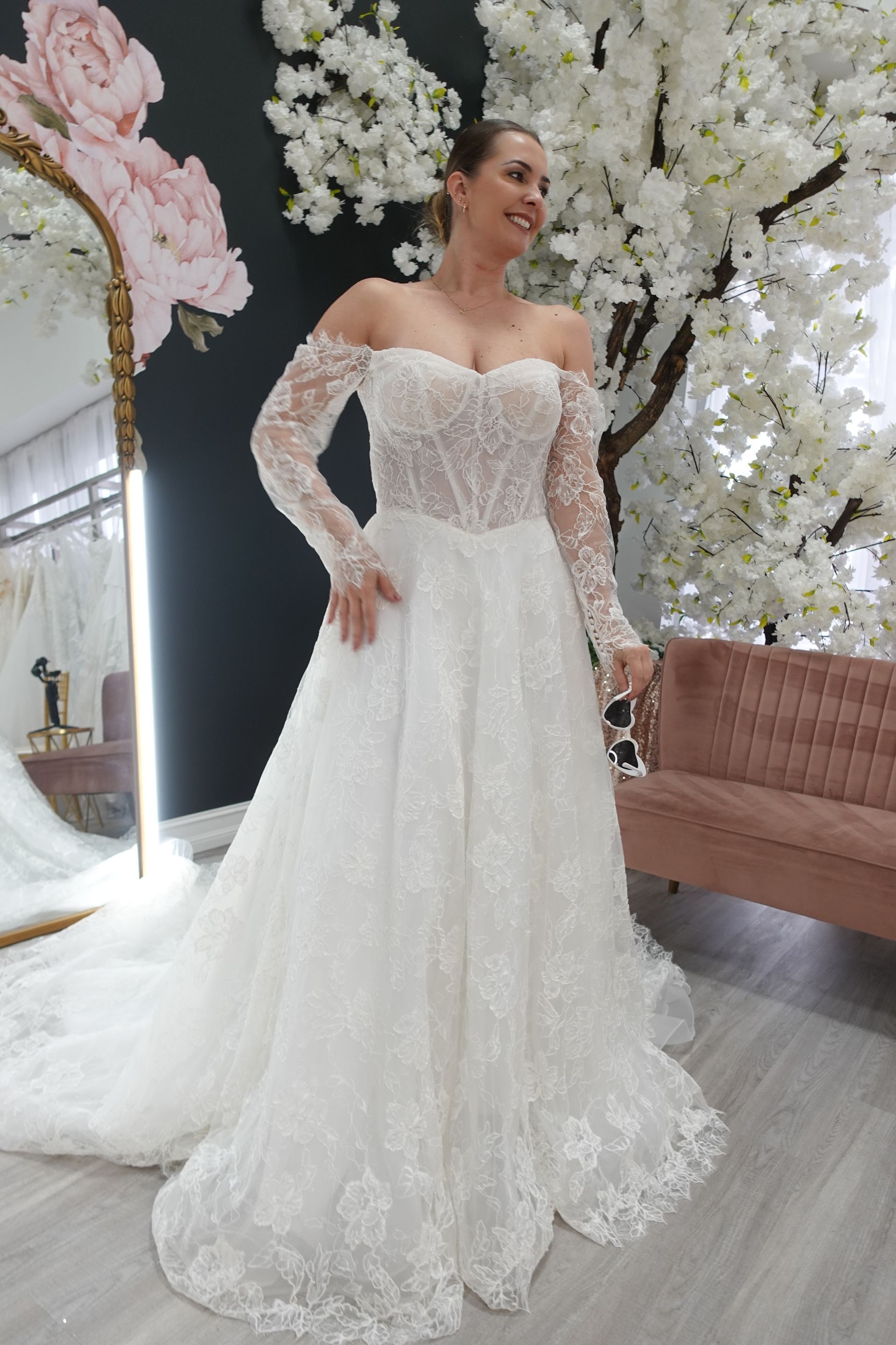 Woman in a white off-the-shoulder wedding gown smiles, posing by a mirror and floral backdrop.