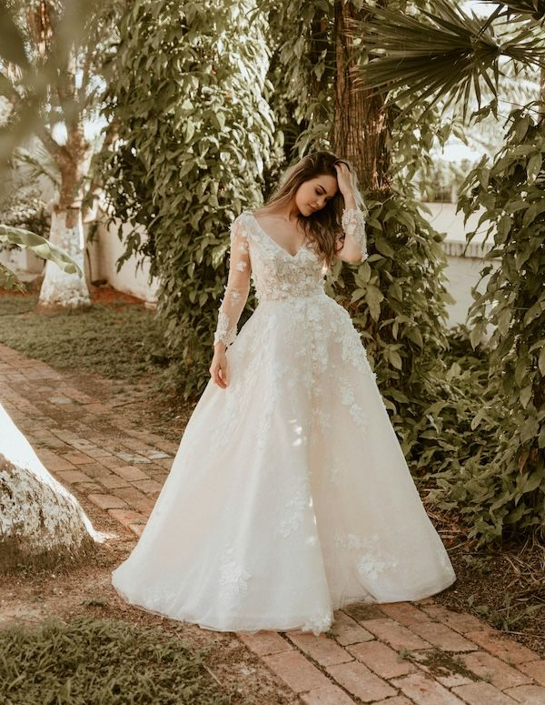 Woman in wedding dress in a garden, hand in hair. She wears a long-sleeved gown with a full skirt.