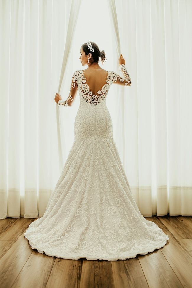 Bride in a white lace wedding dress, holding curtains open, back to the camera, looking at window.