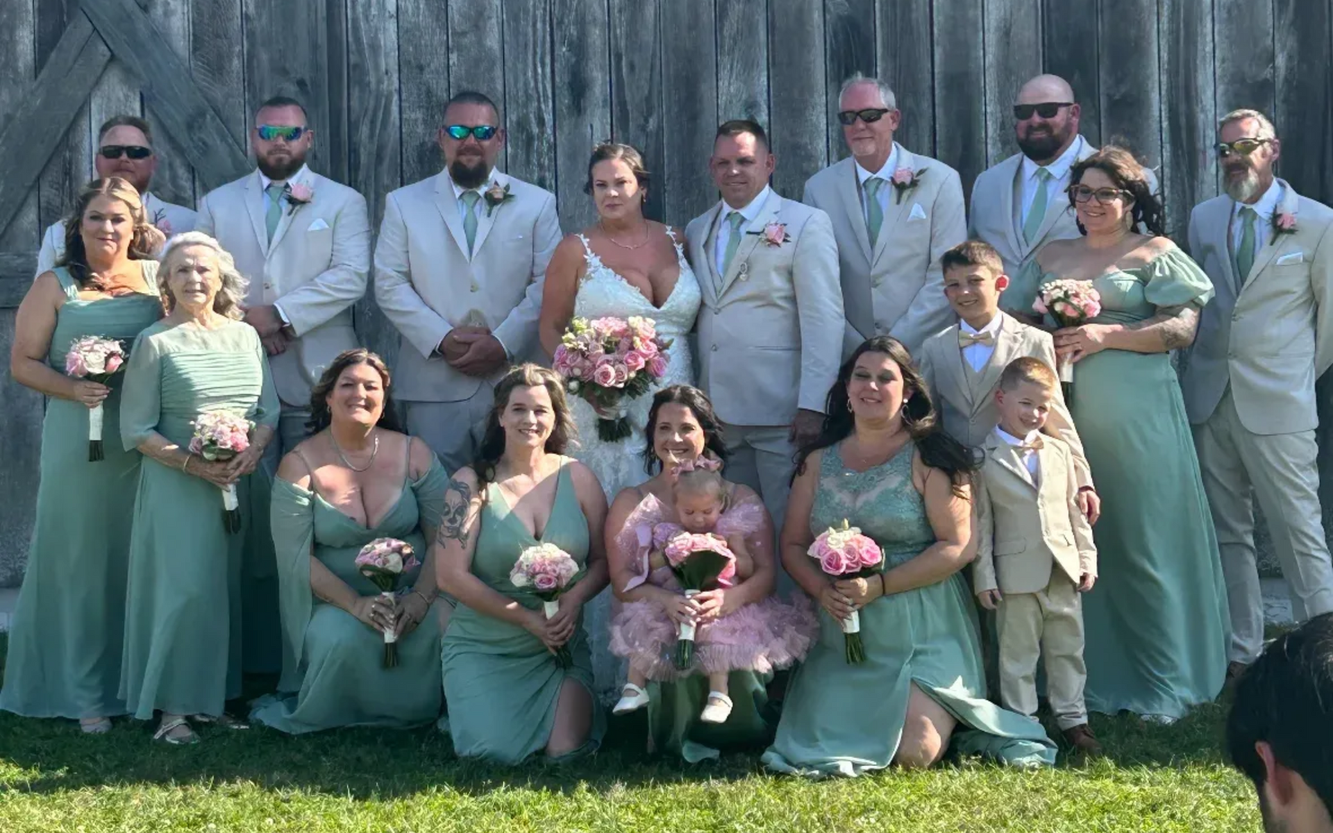 Wedding party posing in front of a barn: Bride and groom with bridesmaids in green, groomsmen in grey suits, and children.