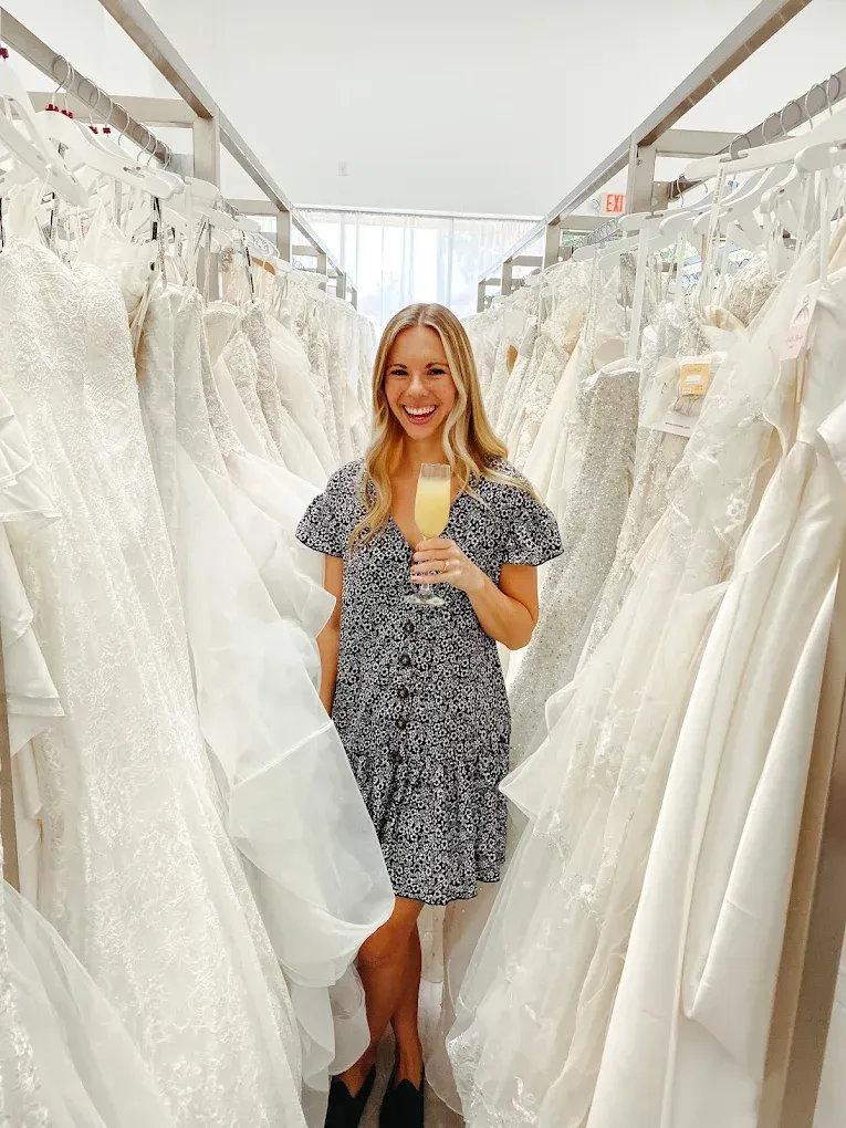 Woman holding a mimosa, smiling in a room filled with wedding dresses.