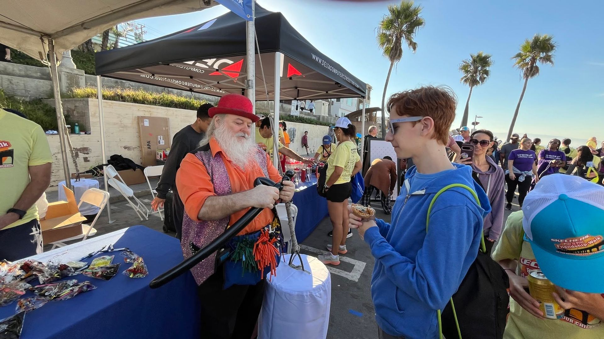 Santa in red hat talks with a boy at an outdoor booth, with people and palm trees in the background.