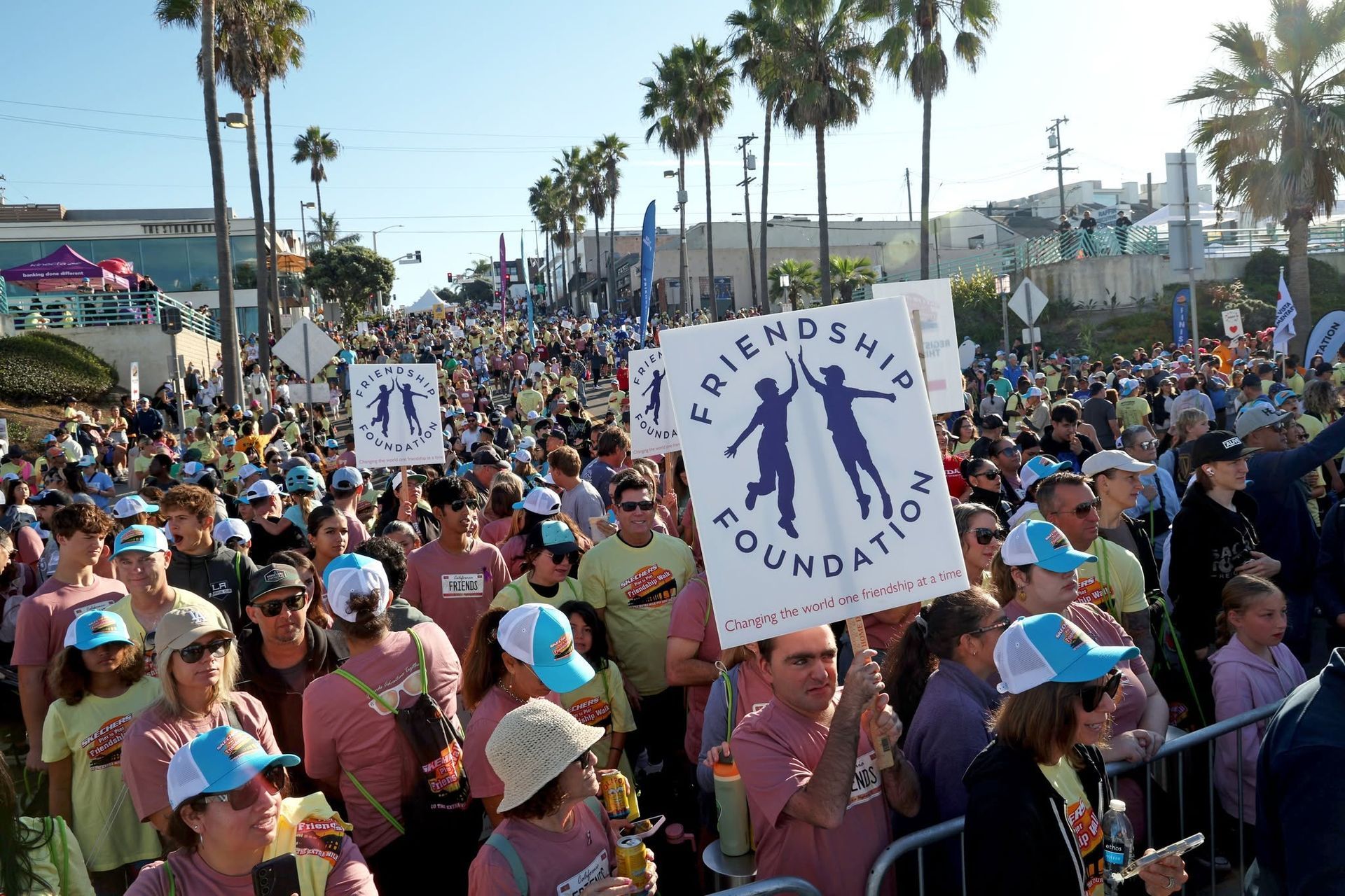 Crowd at a beach walk holding signs for a children’s foundation, with palm trees and bright sun in the background