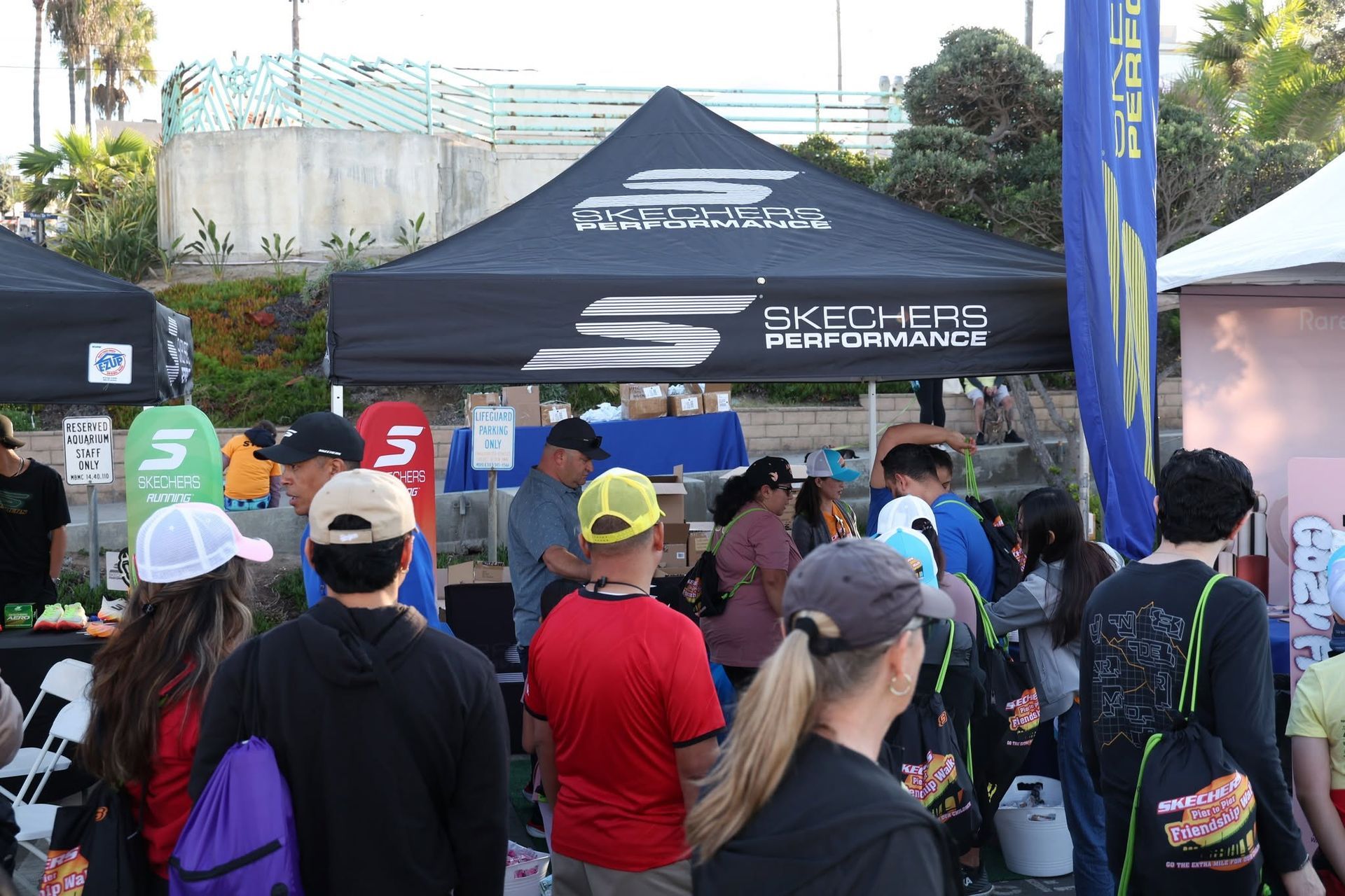 Crowd gathered at an outdoor event booth under a black tent with banners and a performance stage nearby