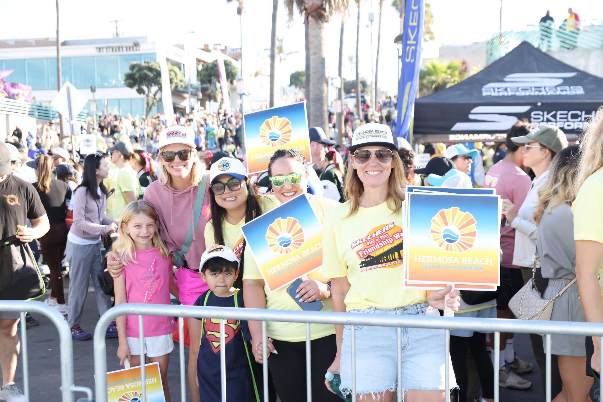 Crowd behind a barricade at an outdoor event, with two people holding colorful signs in the foreground.