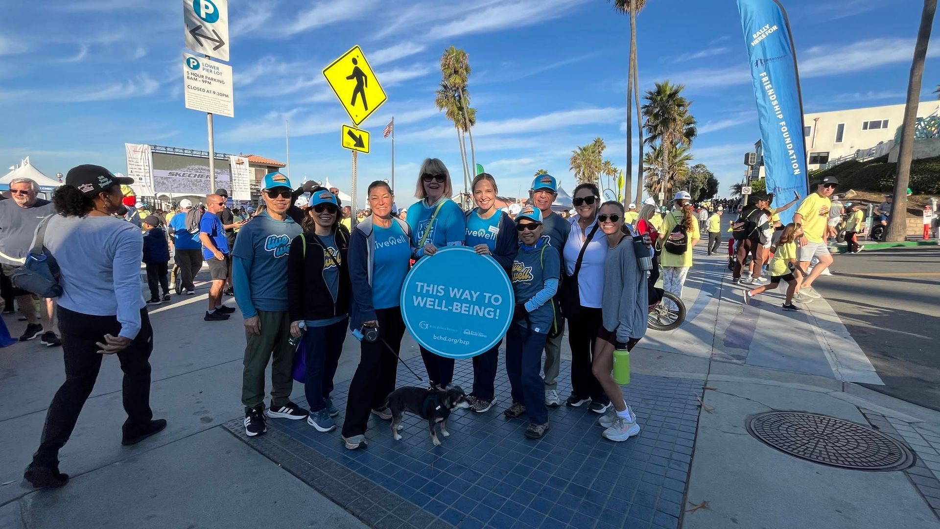 Group of people in blue shirts and masks holding a round sign at a sunny street event.