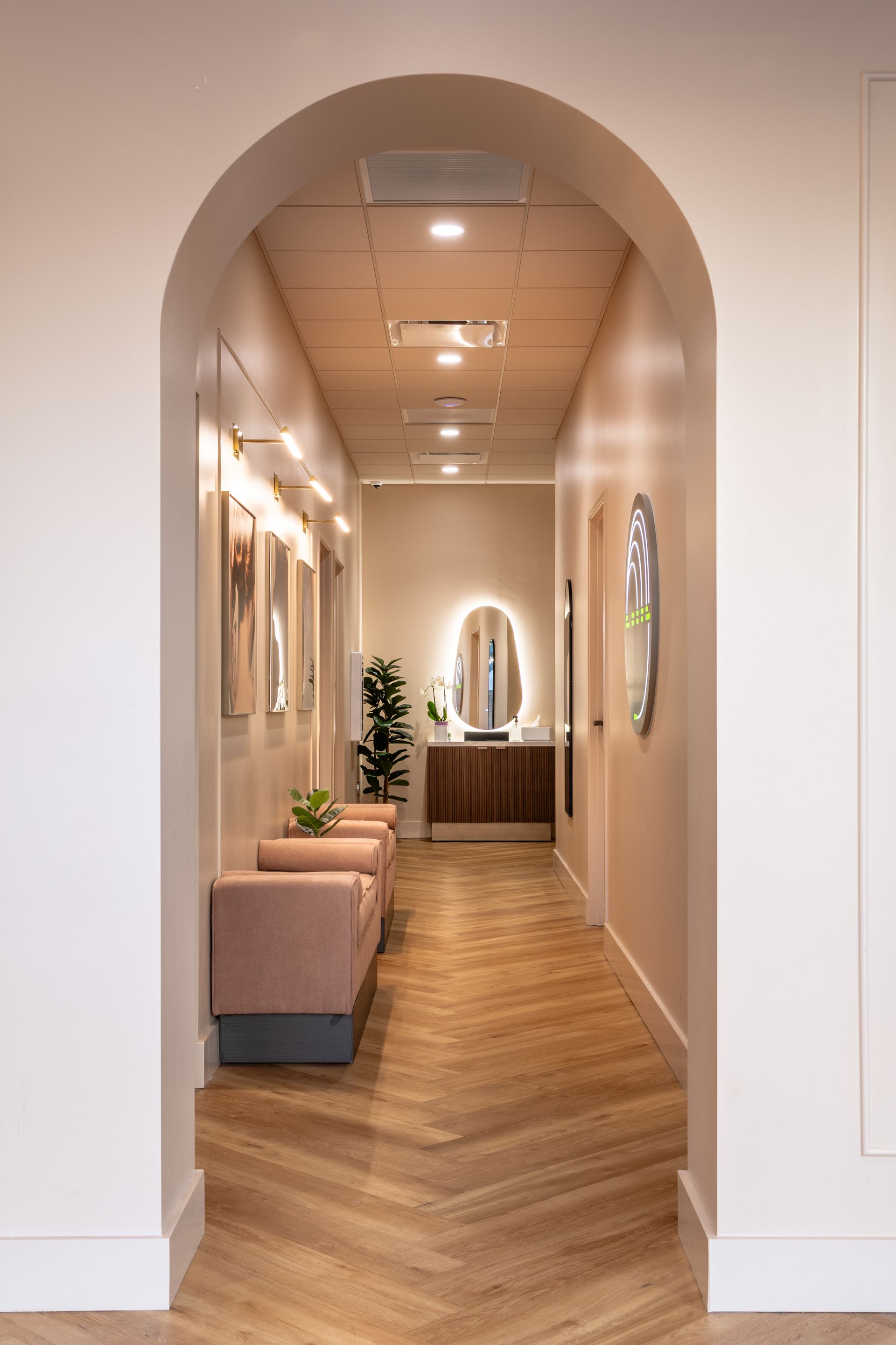 Narrow hallway with arched opening, wood floor, pink bench, wall art, and a lit mirror at the end
