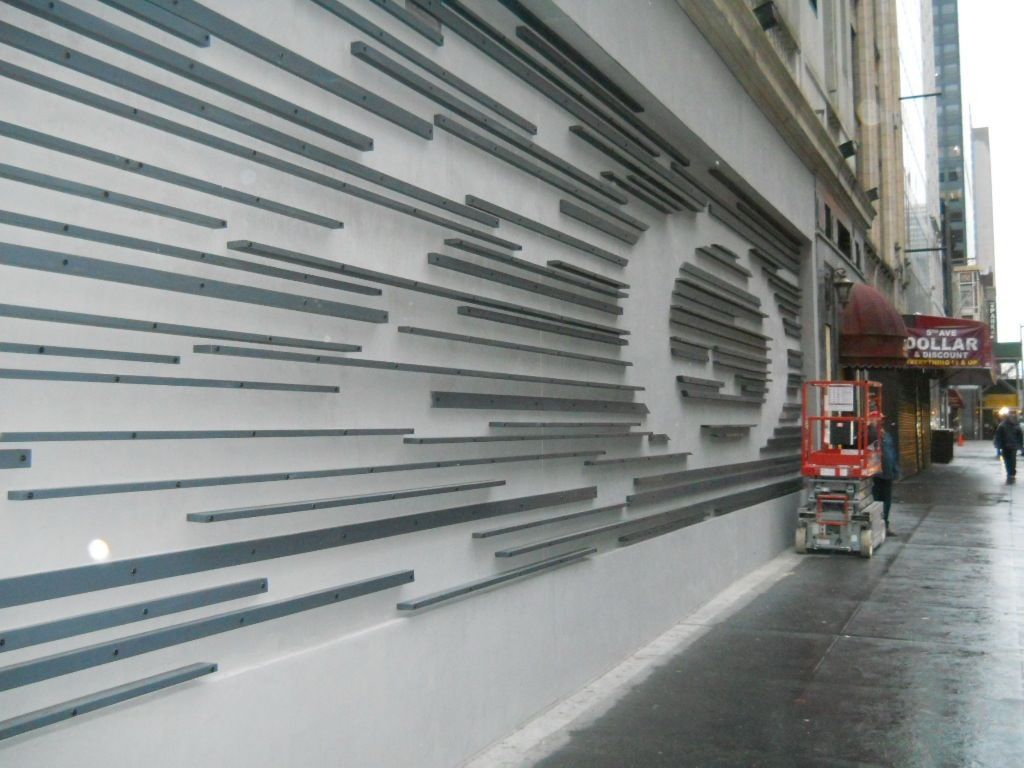 Wet city sidewalk beside a white wall with gray geometric stripes and a small red cart near storefronts