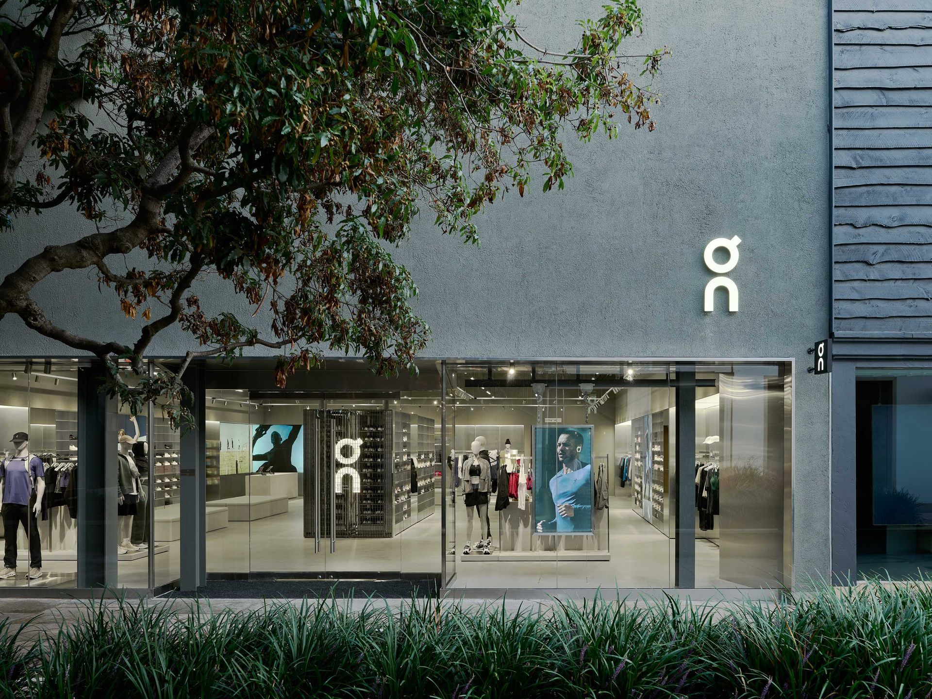 Modern storefront with glass windows, gray facade, and people visible inside under a tree-lined street.