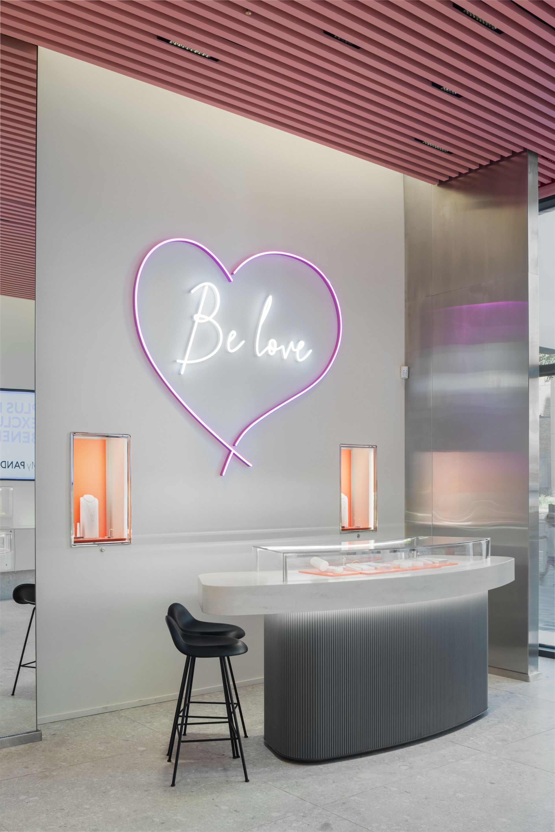 Modern lounge with a neon “Belove” heart sign, curved bar, and tall stools under a pink slatted ceiling