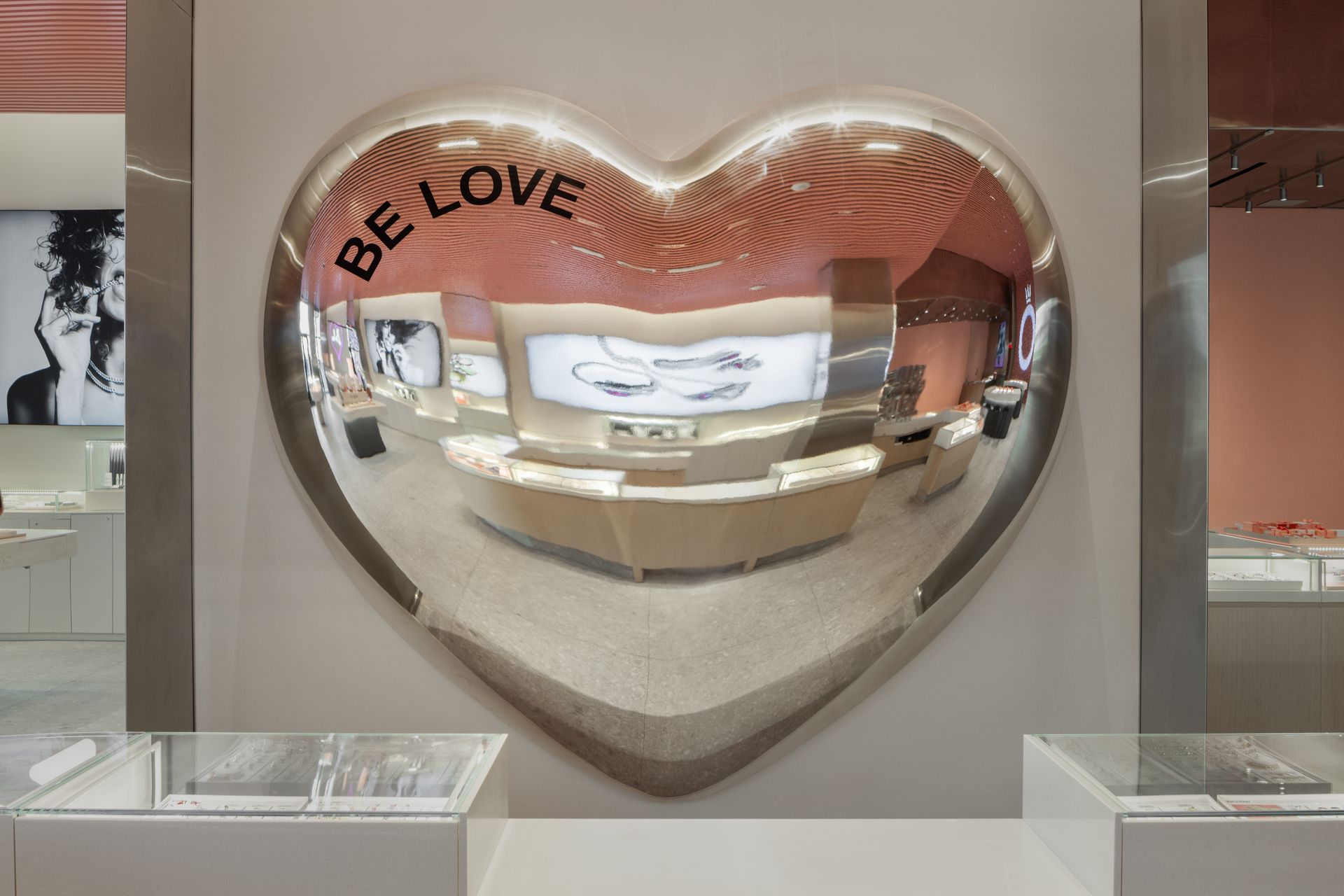 Large mirrored heart sculpture with “BE LOVE” text in a bright gallery display