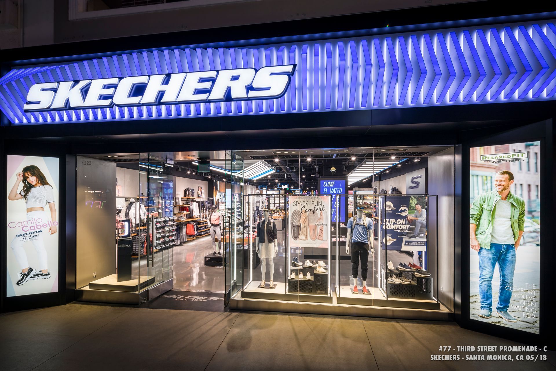 Skechers storefront at night with bright blue sign and glass entrance displaying shoes and clothing.