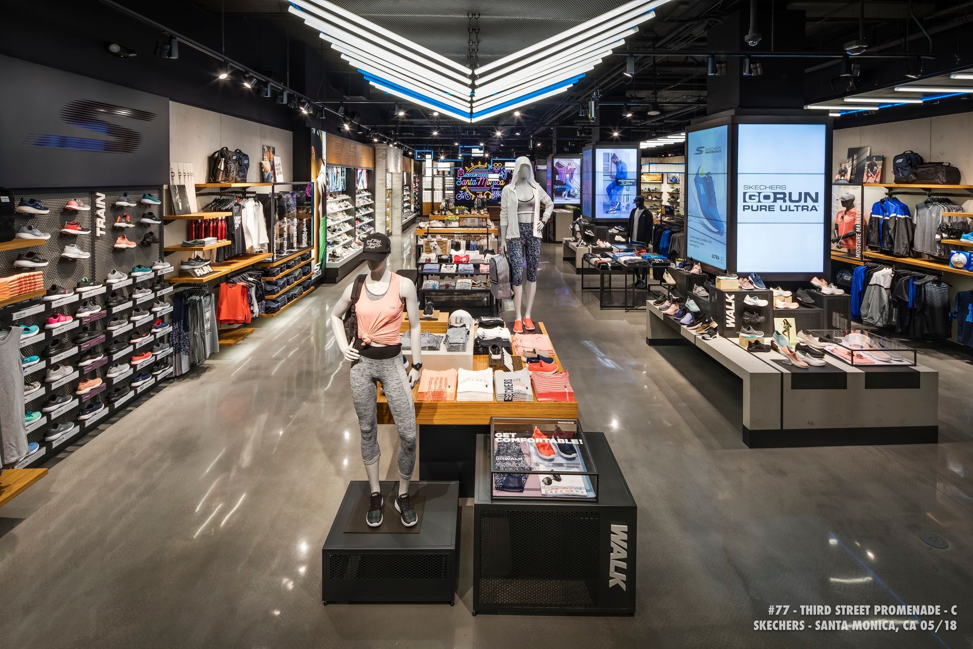 Shoe store interior with sneakers displayed on shelves and tables, and a man standing near the entrance.