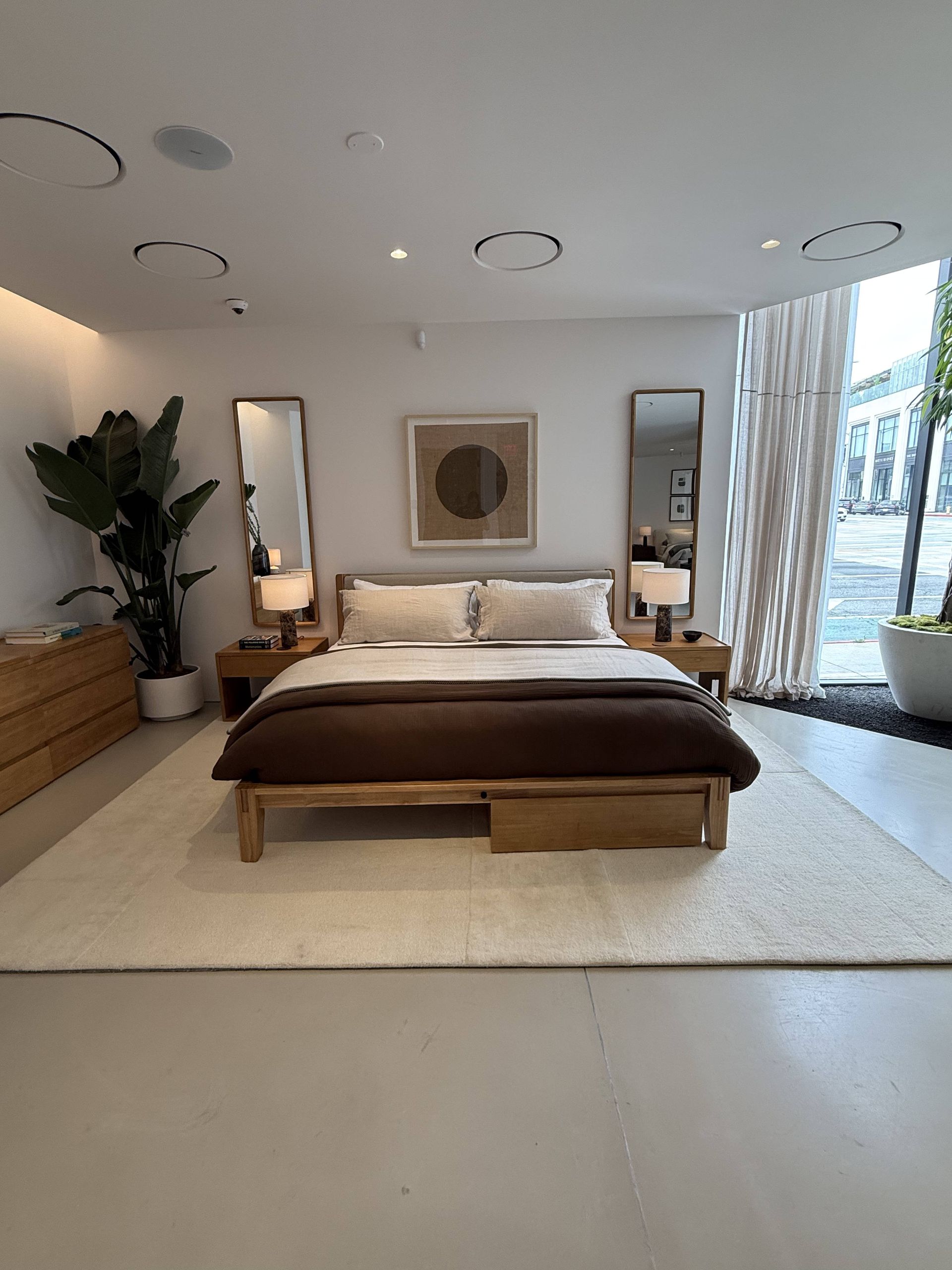 Modern bedroom with a low bed, white rug, wood dresser, and large windows overlooking trees.