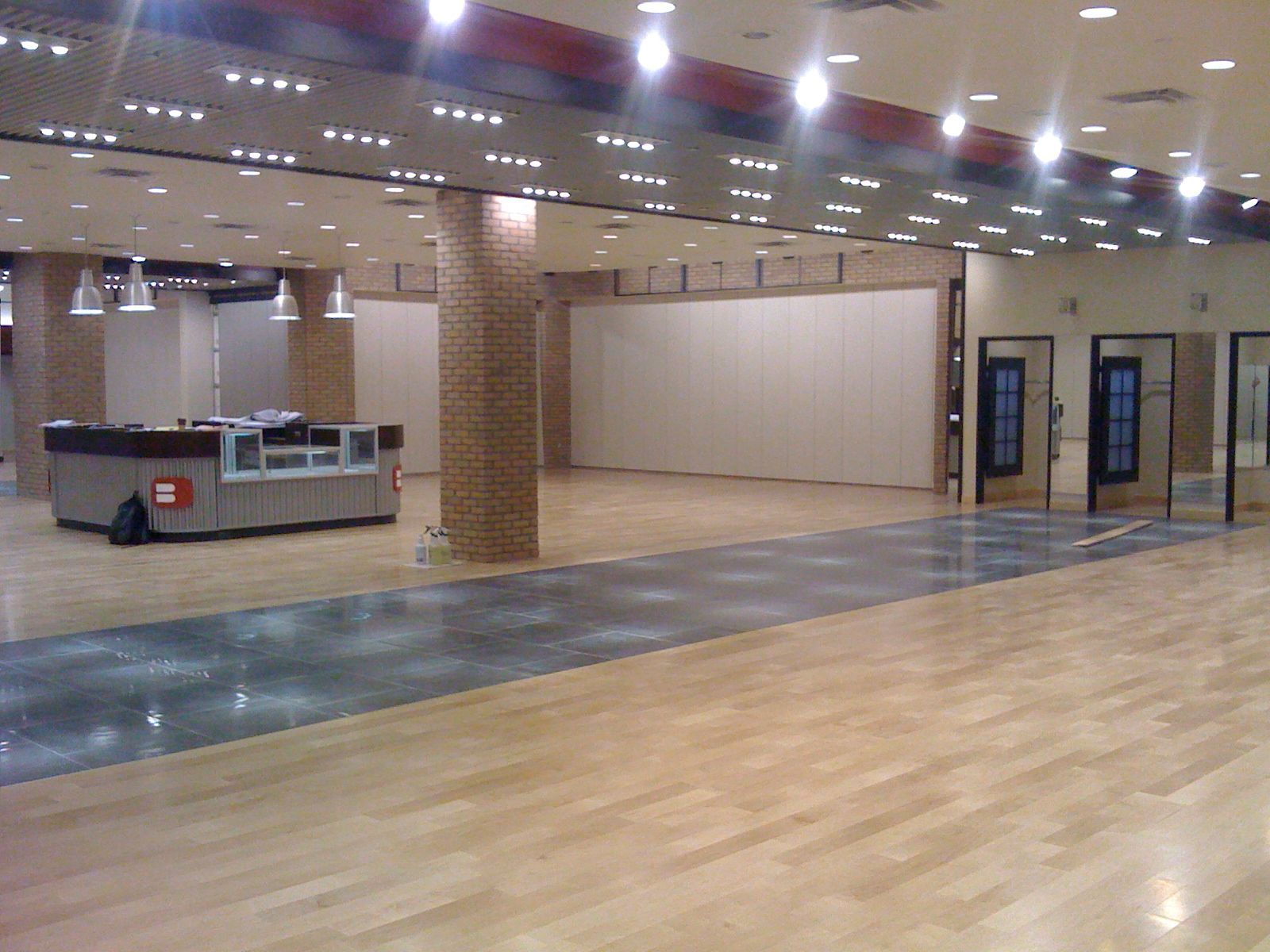 Empty indoor hall with polished wood floor, brick columns, and a kiosk area under bright ceiling lights