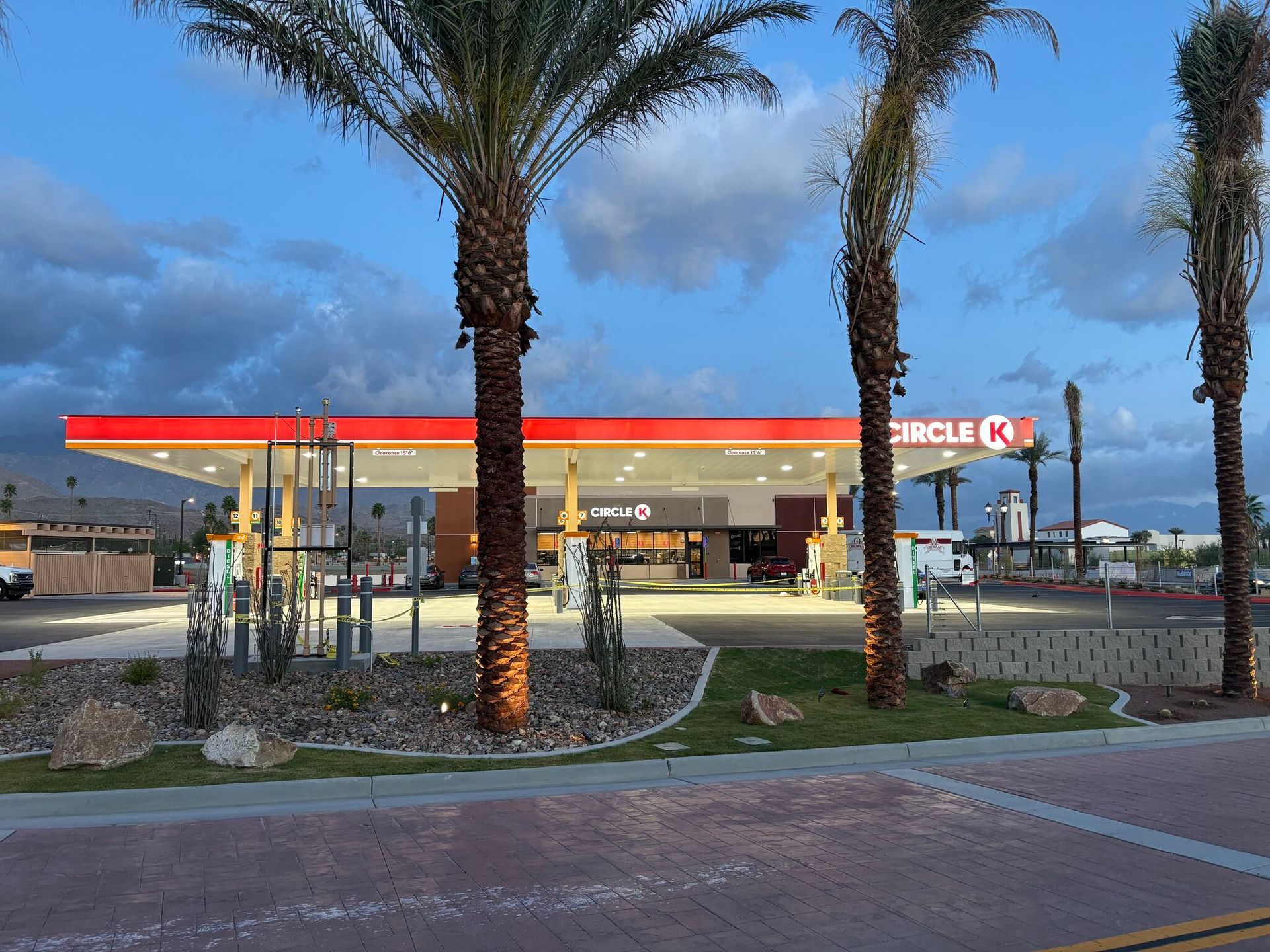 Gas station at dusk with palm trees, red canopy, and parked cars in the lot
