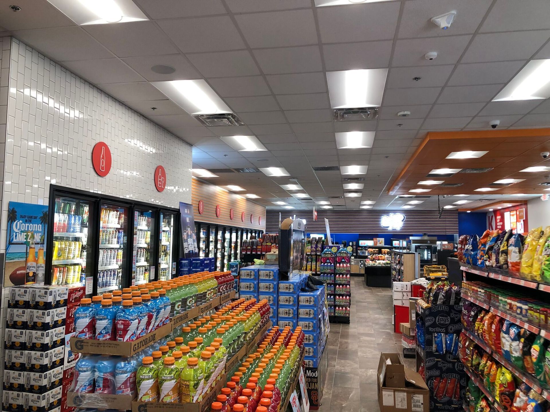 Bright grocery store aisle with refrigerated drinks, snacks, and rows of produce under ceiling lights