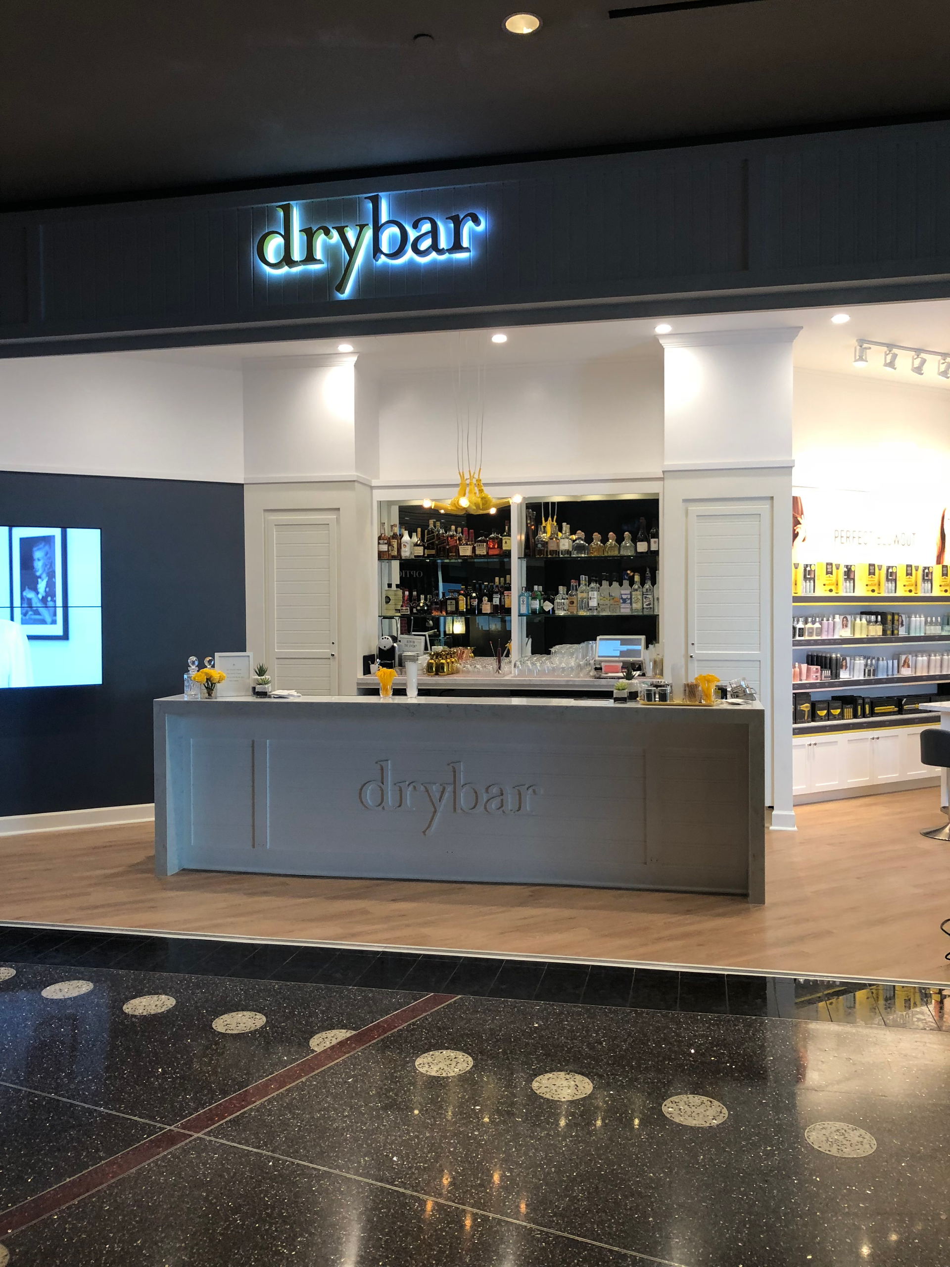 Drybar salon kiosk in a mall, with a lit sign and product displays behind the counter.