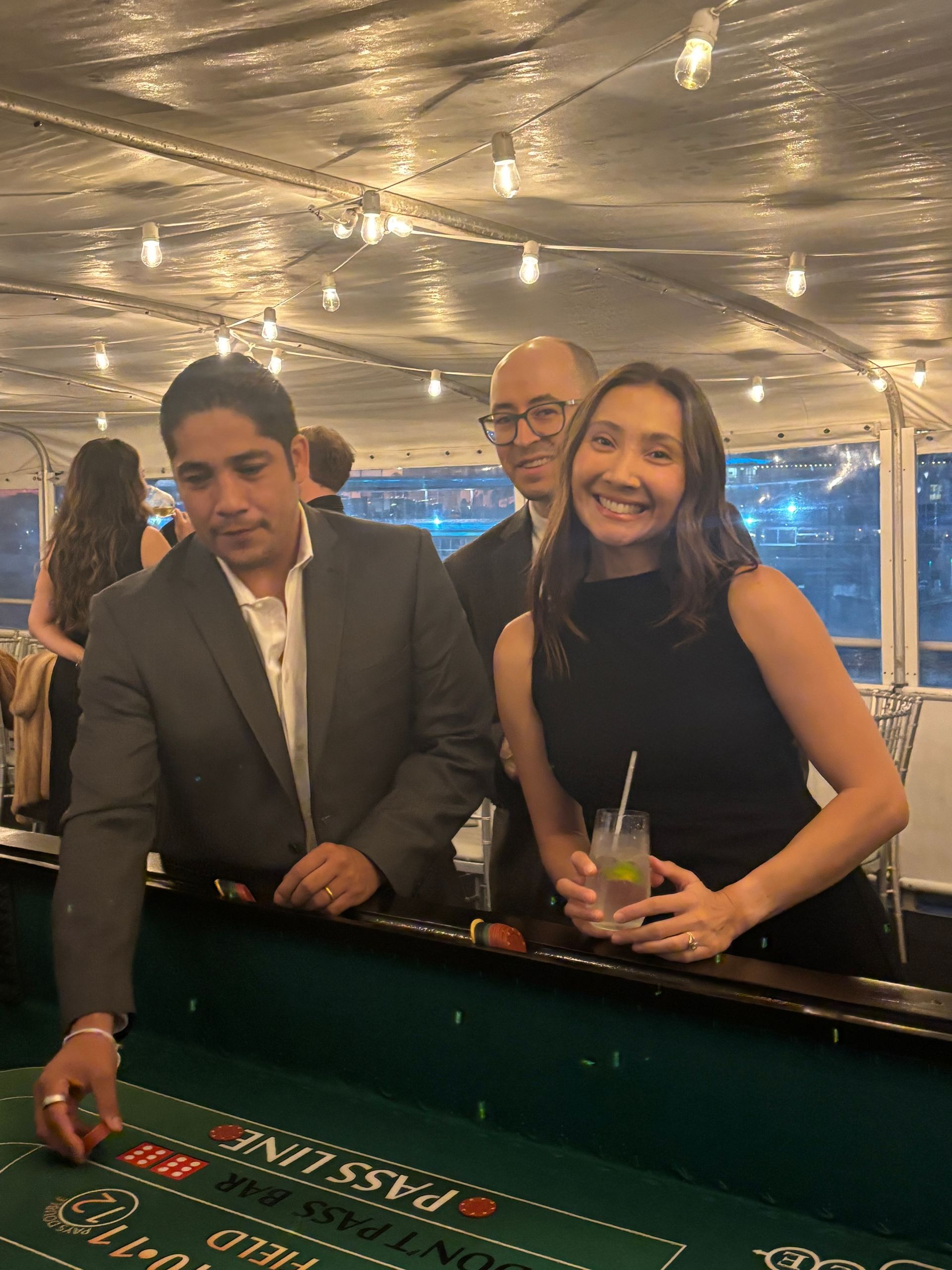 Two people at a casino table under string lights, one smiling and holding a drink.