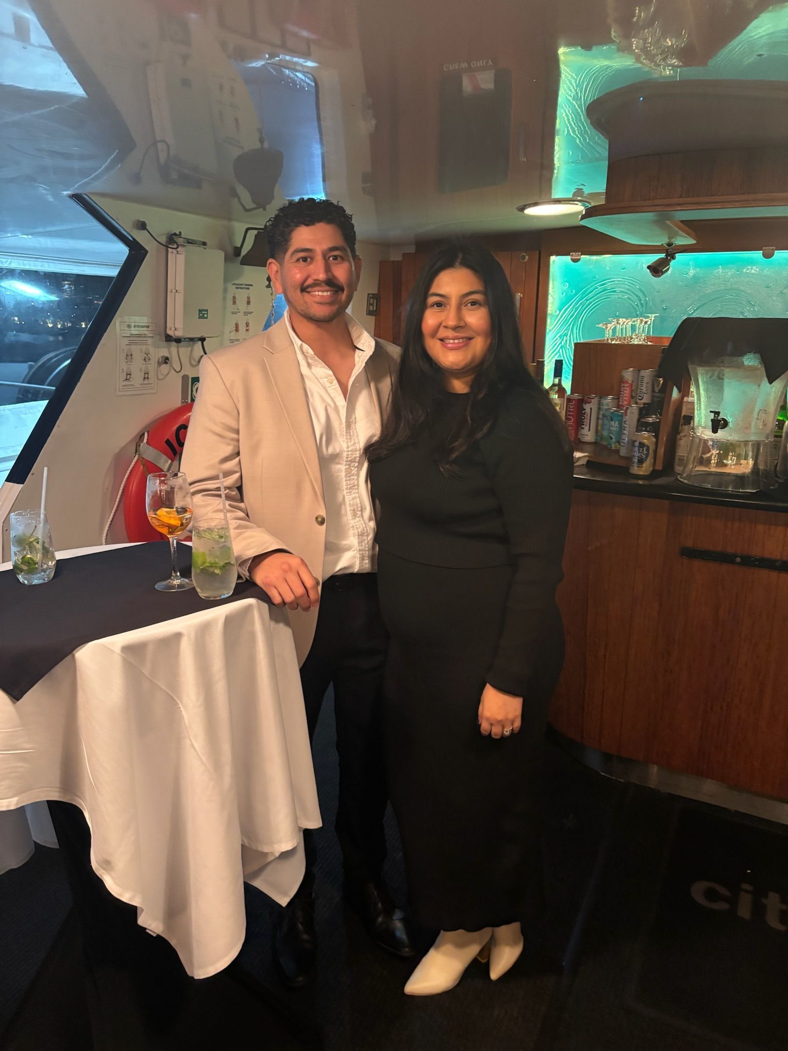 Two people smiling at a bar, standing beside a table with a white tablecloth.