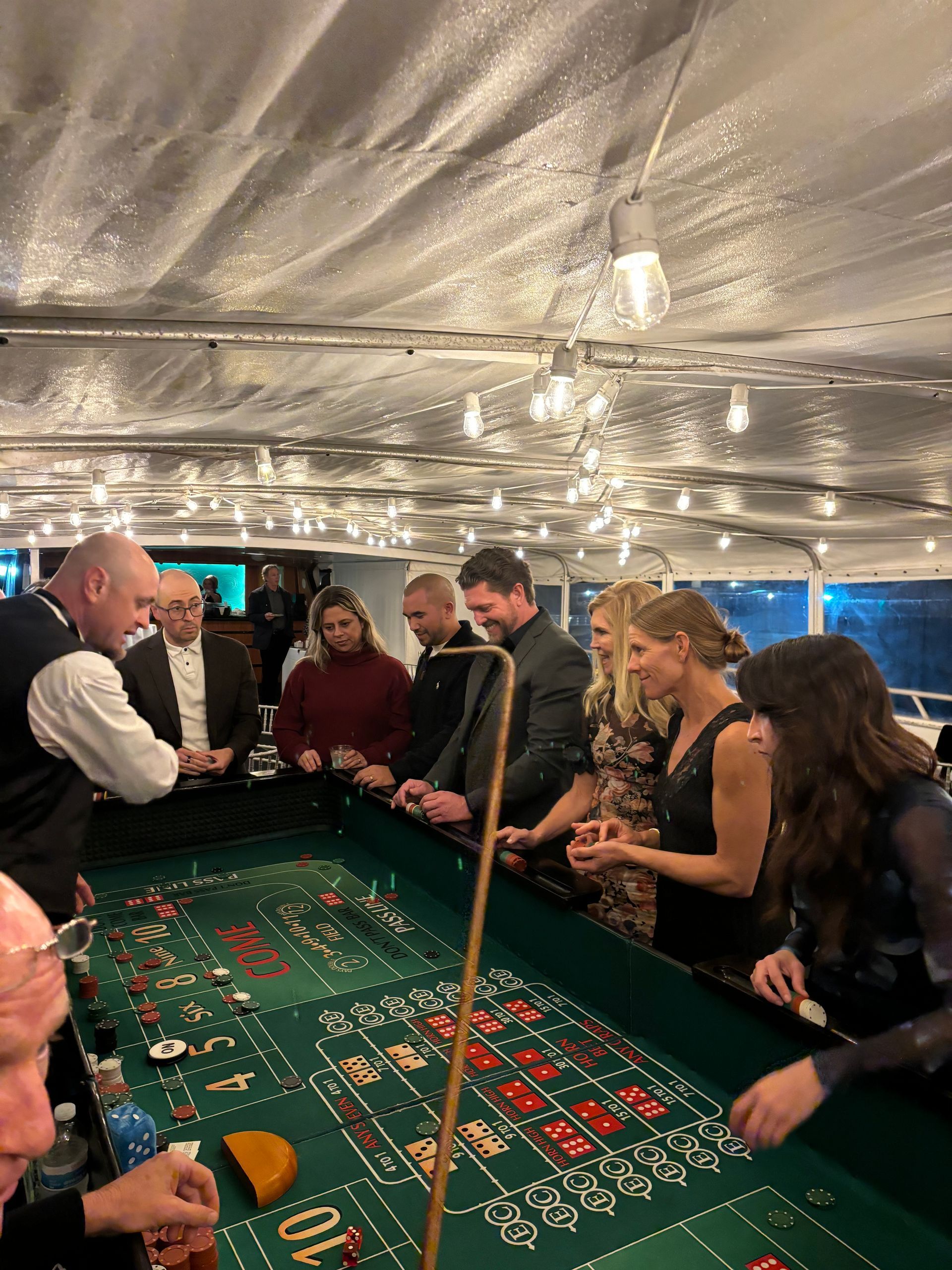 People gathered around a casino table under a white canopy, watching chips and cards on the green felt.